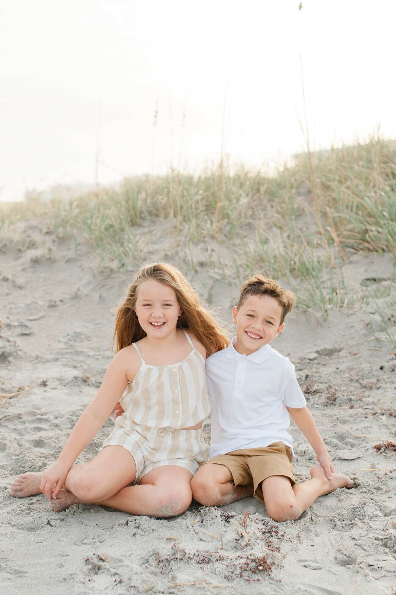 Brother and sister sitting in the dunes smiling at the camera during golden hour