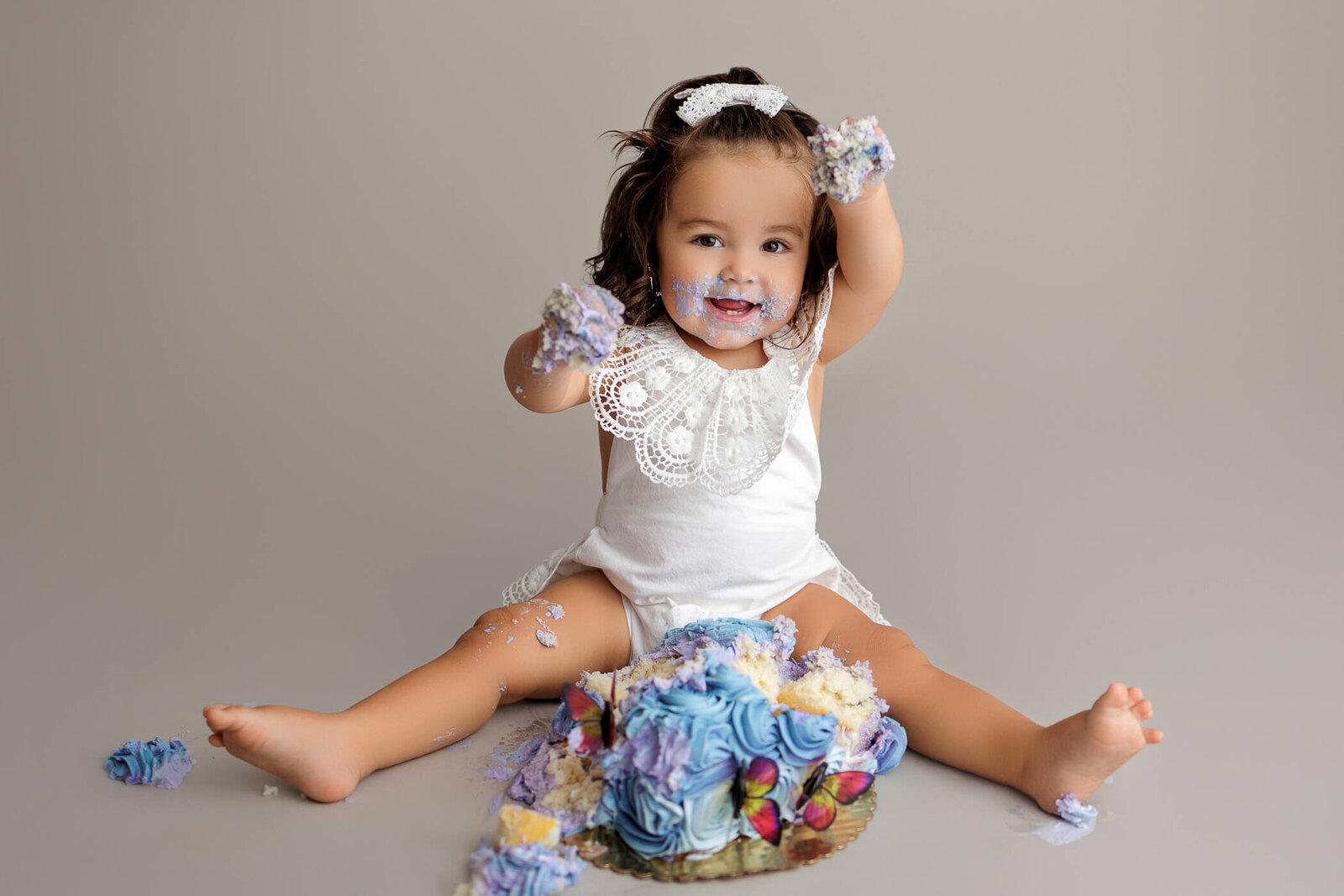 A happy toddler with dark hair, wearing a white lace romper and a white headband, sits on a gray backdrop. The child is joyfully smashing a colorful cake with purple, blue, and yellow frosting, with frosting smeared on their face and hands. The toddler raises both hands in the air, covered in cake, creating a fun and celebratory atmosphere.