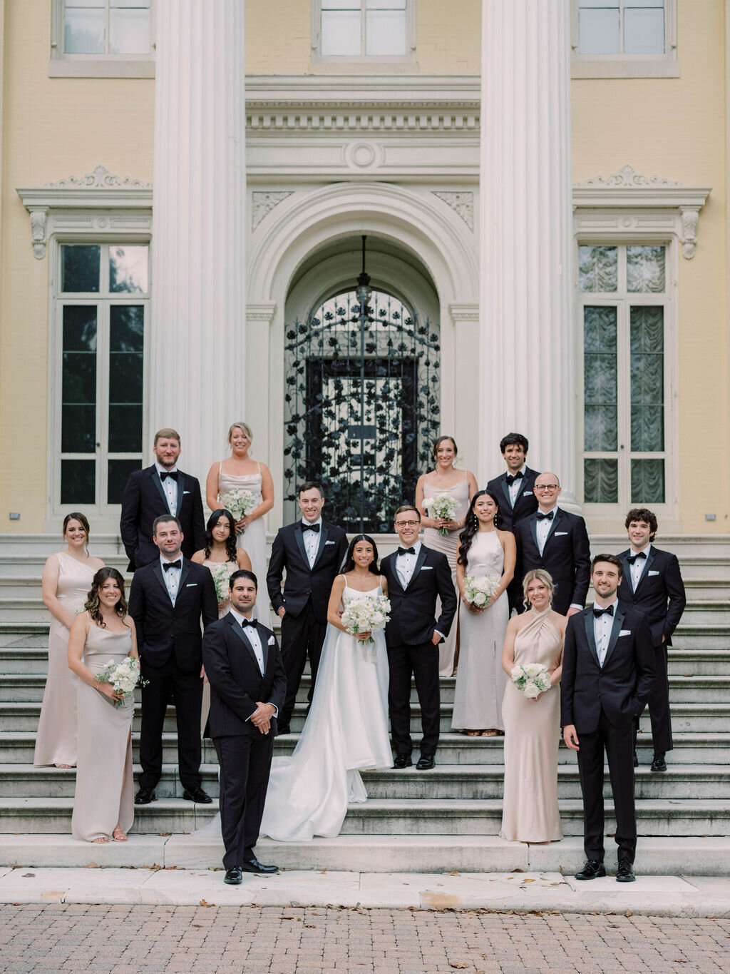 The black tie bridal party standing on the stairs with the bridesmaids wearing champagne-colored silk slip dresses and groomsmen wearing black tuxedos.