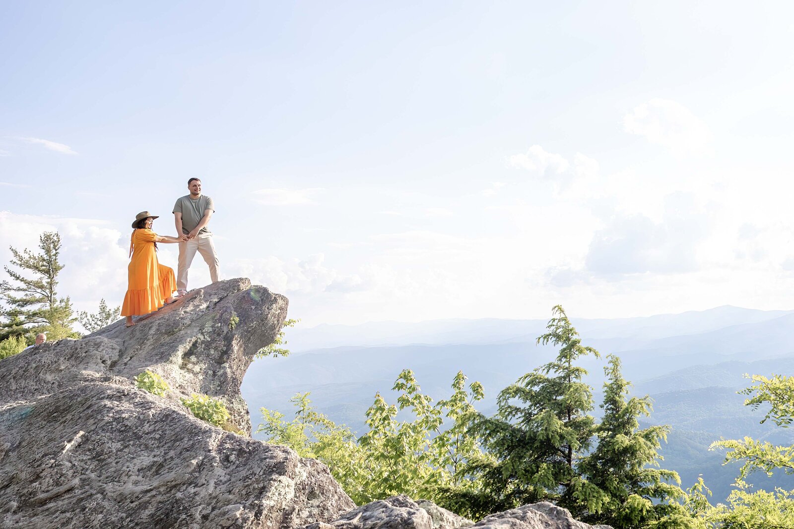 An adventurous couple stands on a dramatic mountain cliff, The Blowing Rock, with the man helping the woman, who wears a flowing orange dress, balance gracefully. The expansive view of the rolling mountains and bright sky emphasizes the adventurous spirit of North Carolina wedding photography. 