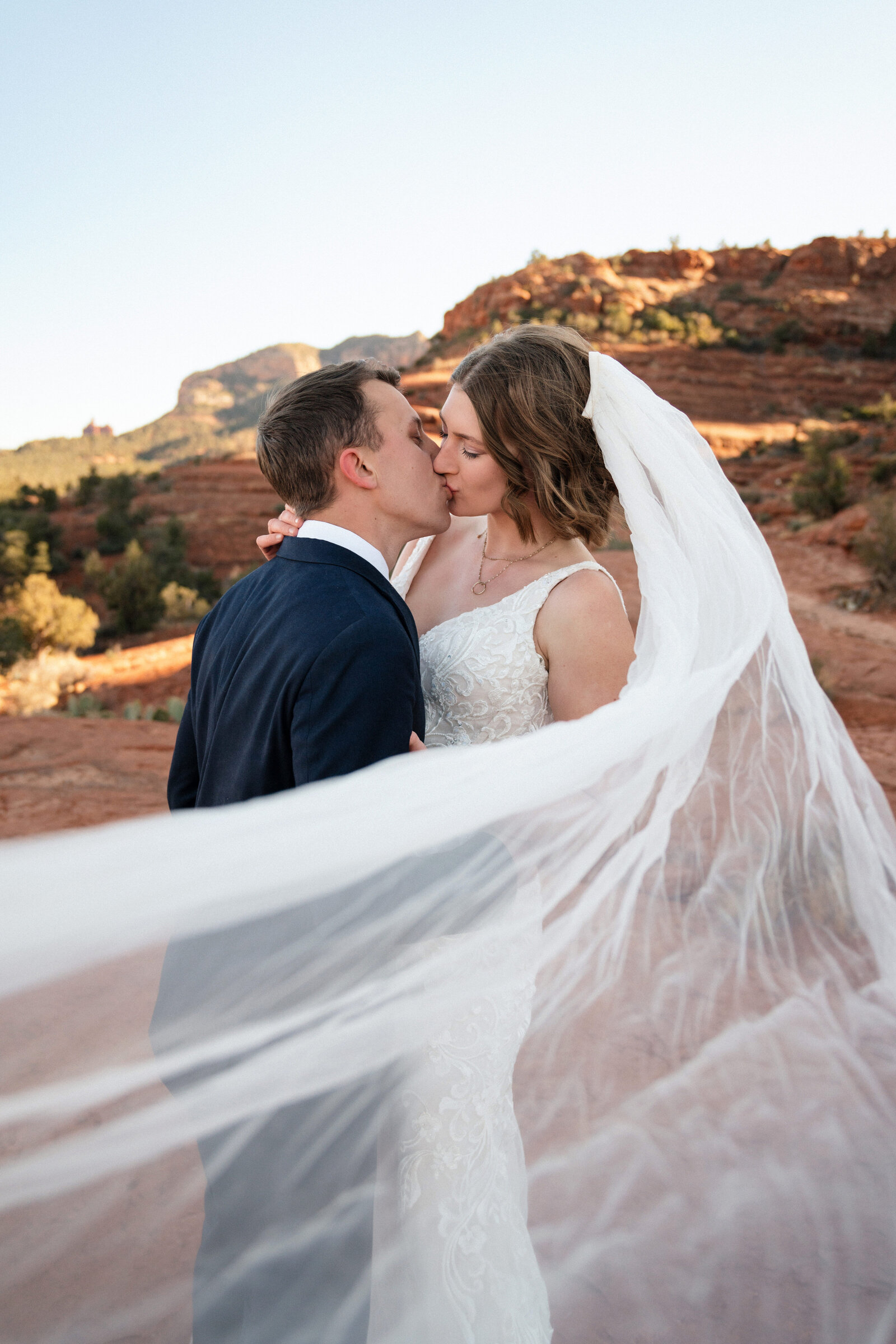 Bride and groom sharing a romantic kiss with the bride’s veil flowing in the wind during their elopement at Cathedral Rock in Sedona, Arizona, with stunning red rock formations in the background.