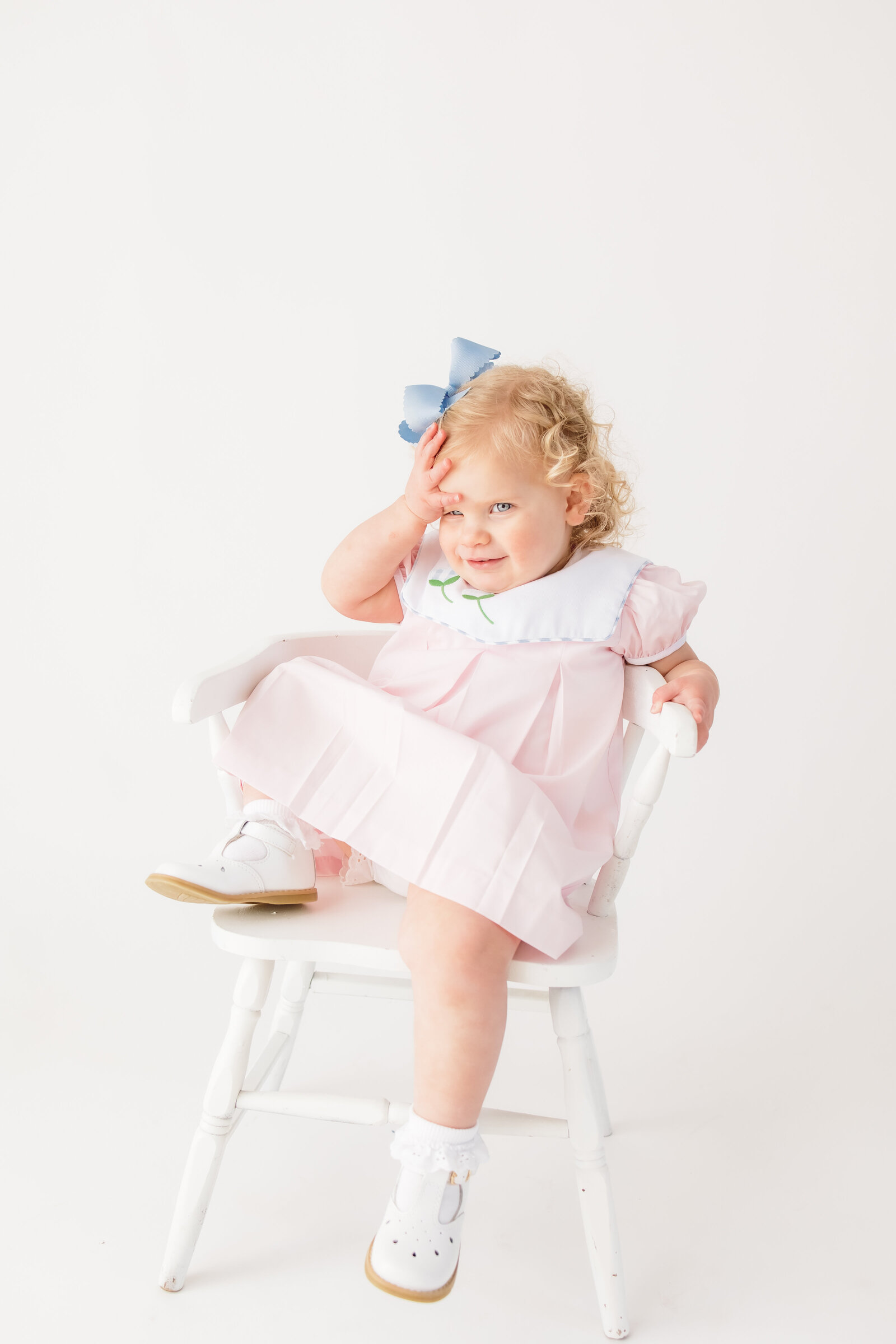 Little girl with her hand on her head  lounging on a white chair in front of a white back drop
