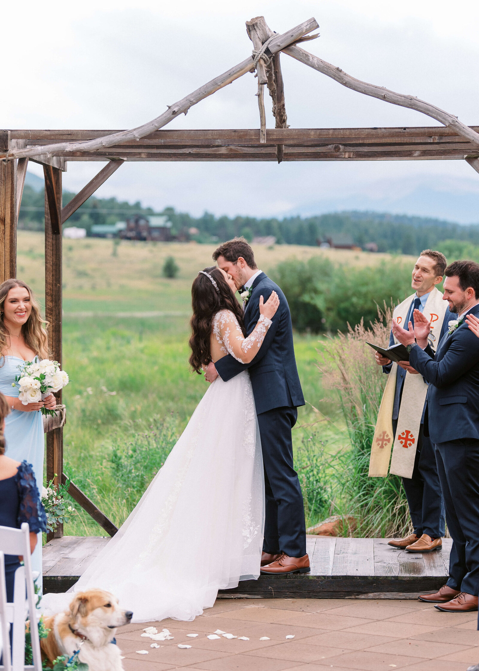 groom and bride kissing at the alter with the mountains in the background and wedding party clapping
