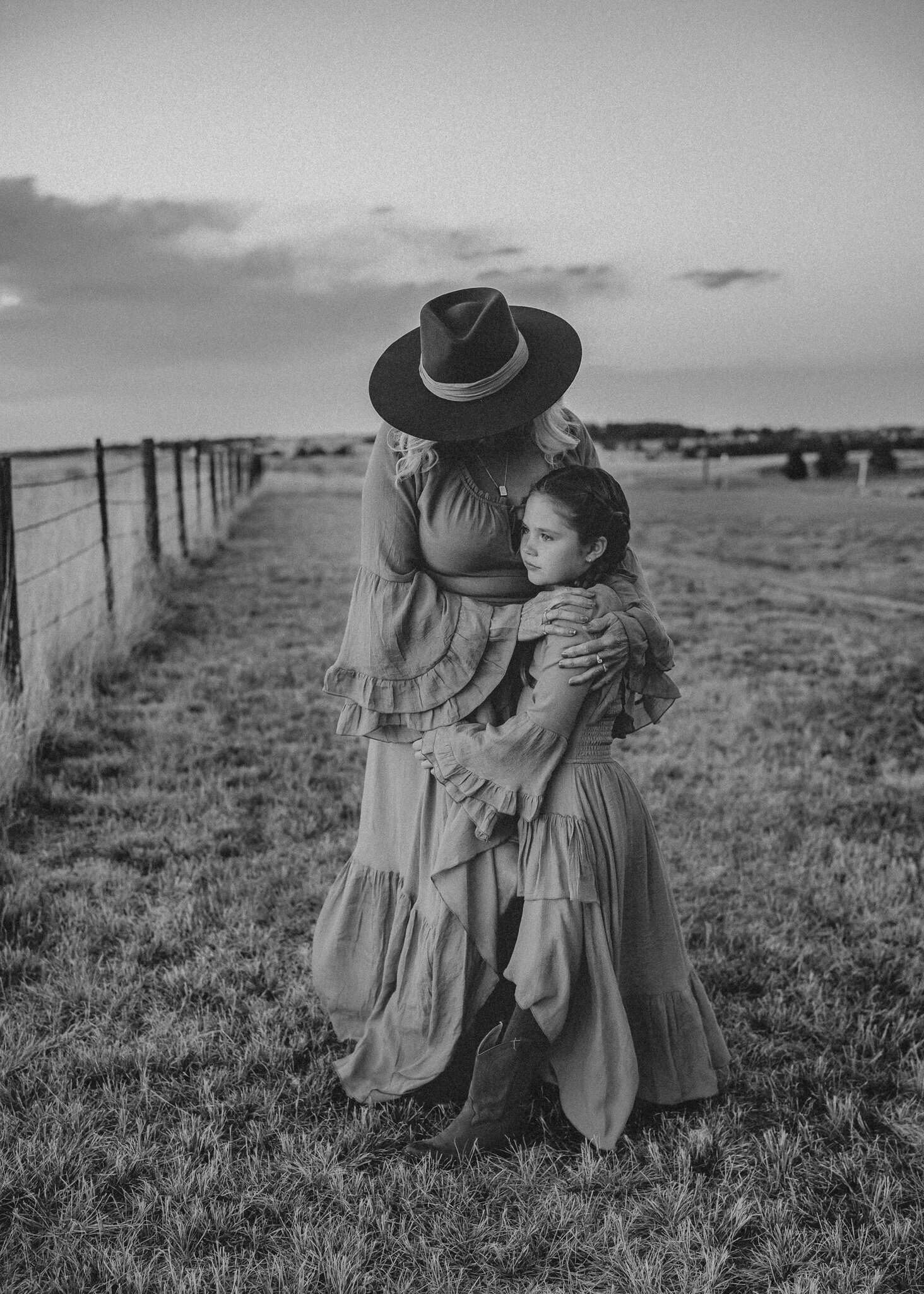 During a grandparent session, a granddaughter hugs, her grandmother in an open field setting as they both look at the beautiful sunset.