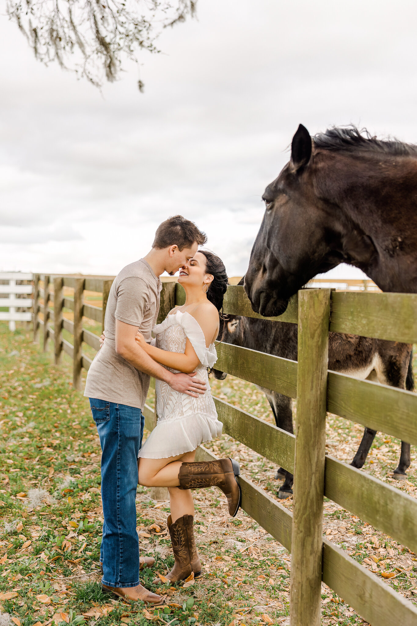 A woman is against a fence almost kissing a man as a horse looks at them