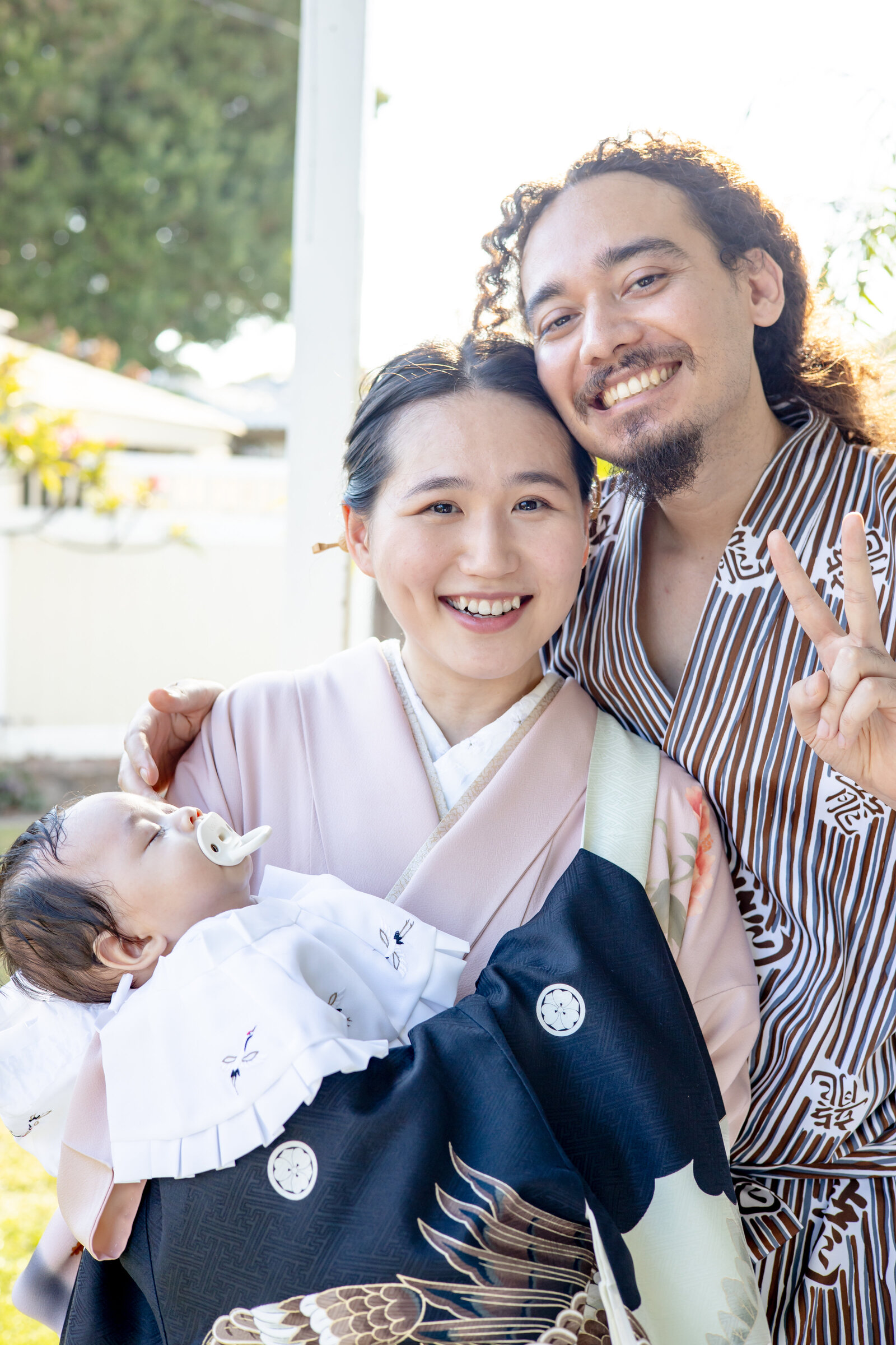 Mother in pale pink kimono holding baby in black celebratory kimono with smiling partner making a peace sign | 初参り 家族写真 ロサンゼルス ピンク着物のお母さんと黒祝い着の赤ちゃん パートナーはピースサイン