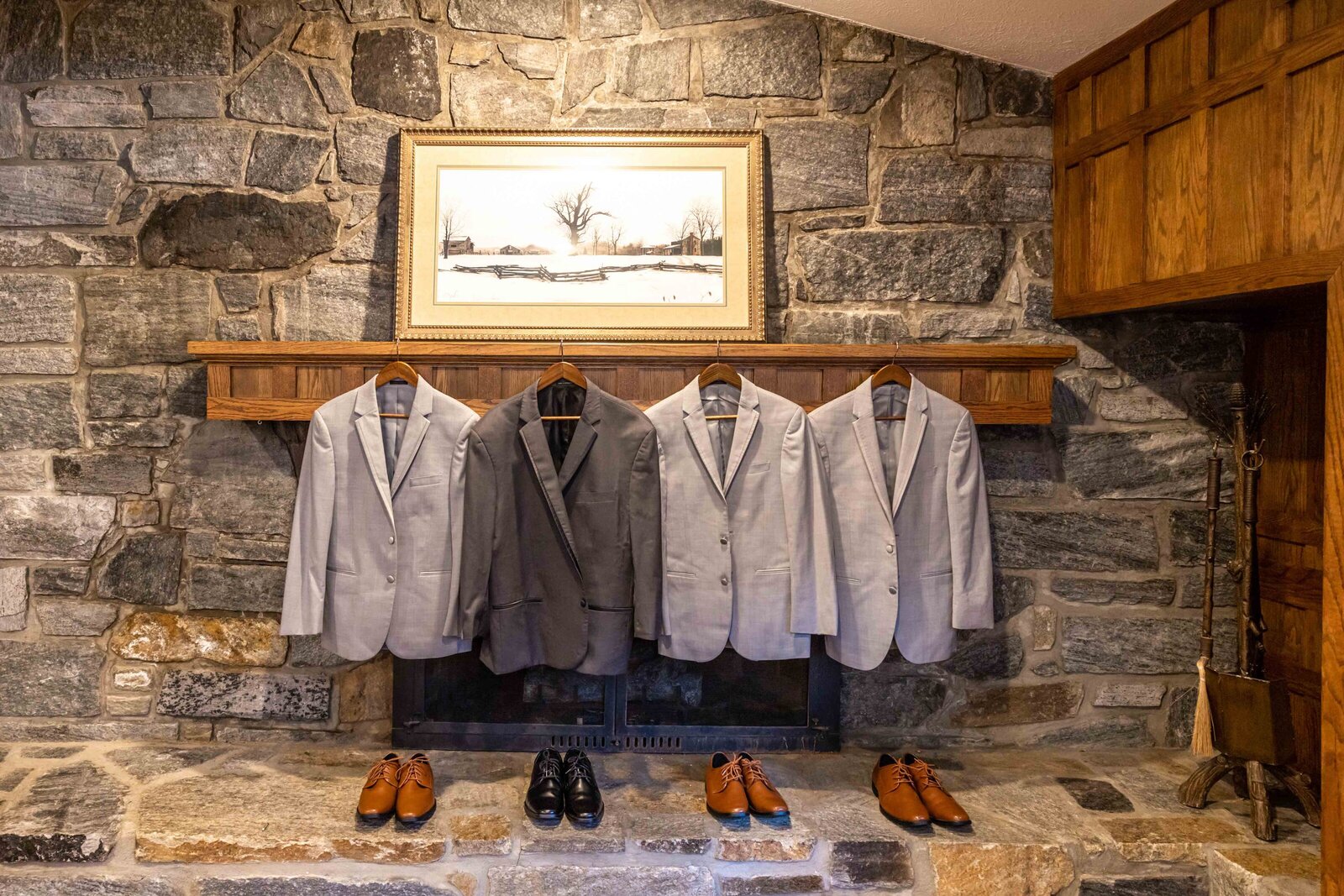 A display of groom and groomsmen suits in shades of gray and brown leather shoes arranged on a stone fireplace mantel, captured during a rustic Boone, NC wedding, highlighting classic and elegant wedding style.
