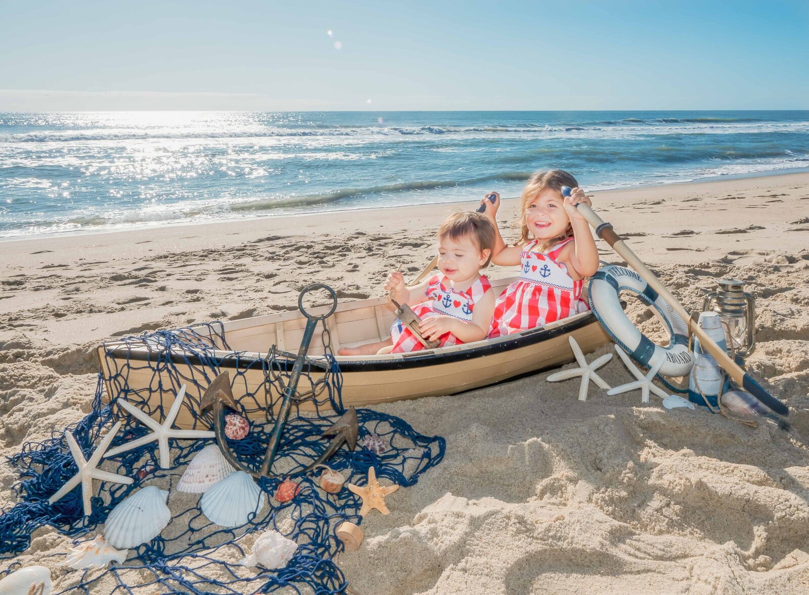 Two young children sit in a decorative boat on the sandy beach, surrounded by seashells and nautical props. Dressed in matching red and white outfits, they enjoy a sunny day by the ocean. This image highlights artistic family photography in Wilmington, NC, capturing whimsical childhood moments.