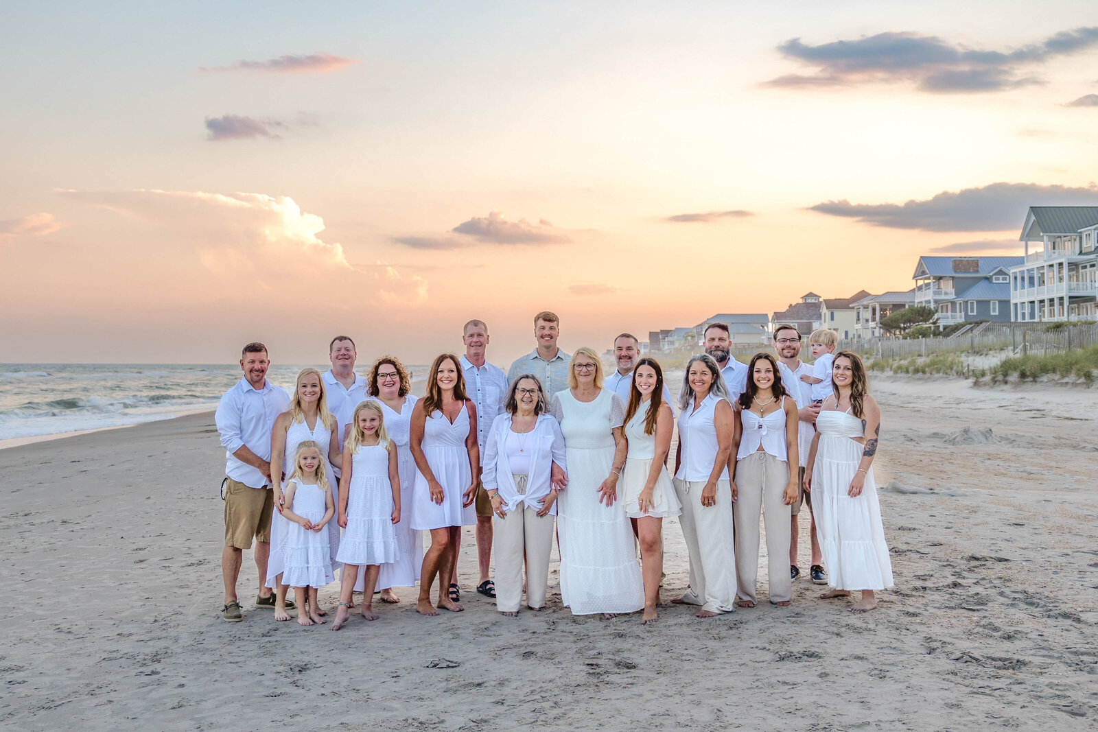 A large family dressed in white poses on the beach at sunset, with coastal homes in the background. The soft evening light creates a serene atmosphere, perfect for North Carolina family photography, capturing elegant and joyful family gatherings.