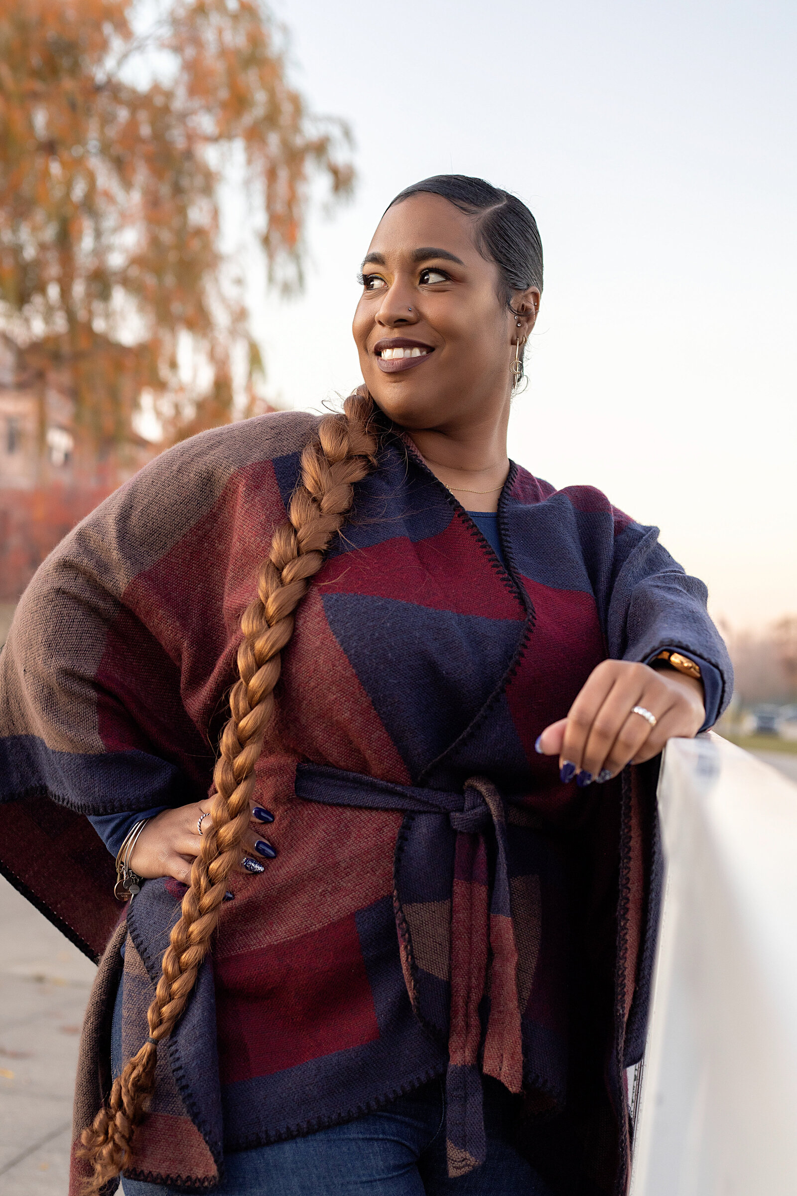 African American woman in red and blue sweater jacket, blue jeans, and a log braid posing by a rail