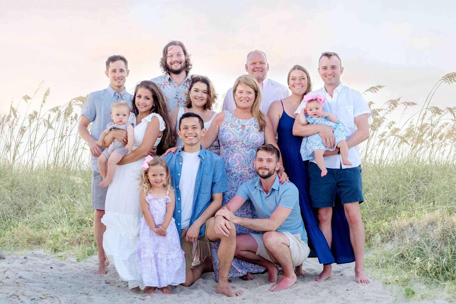 A large, cheerful family gathers on the sandy shores of Carolina Beach, dressed in pastel colors. The group includes parents, children, and grandchildren, all smiling warmly amidst the beach grass and soft evening light. Perfect for those looking for professional family photography in NC.