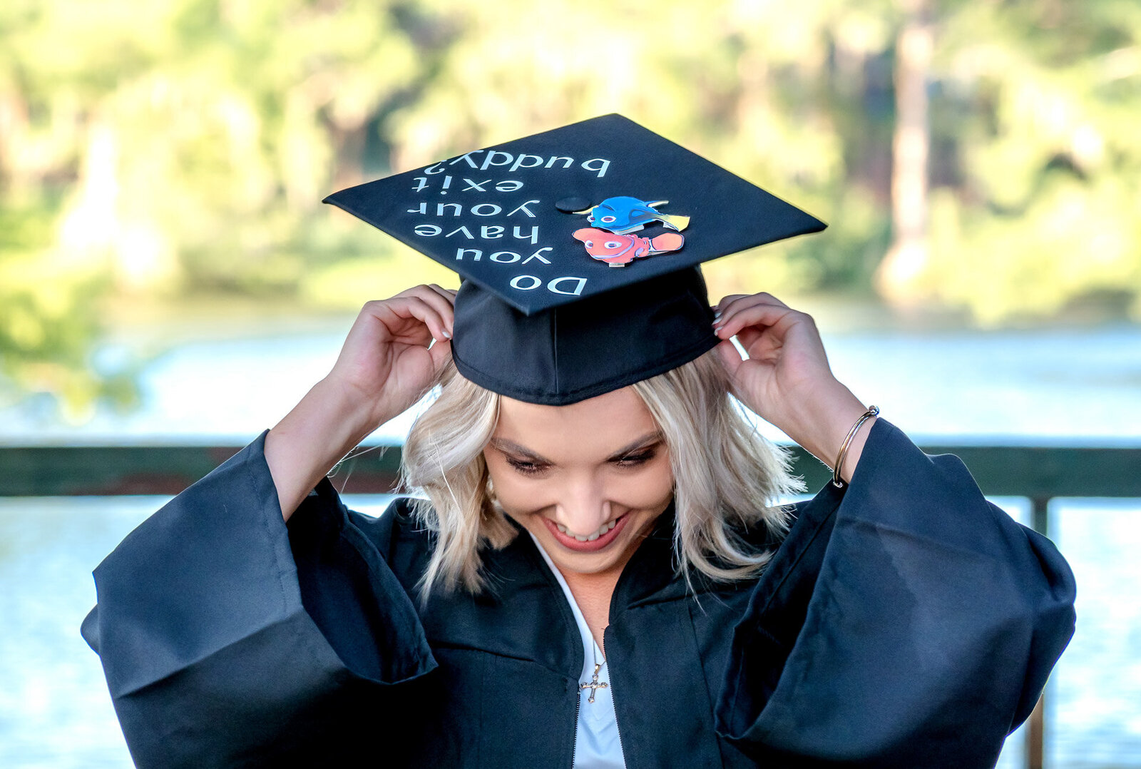 A CFCC graduating senior in a cap and gown, smiling while adjusting her decorated cap at a scenic location in Wilmington, NC. This photo highlights the joy of graduation, perfect for those seeking senior photographer services in Wilmington, NC.