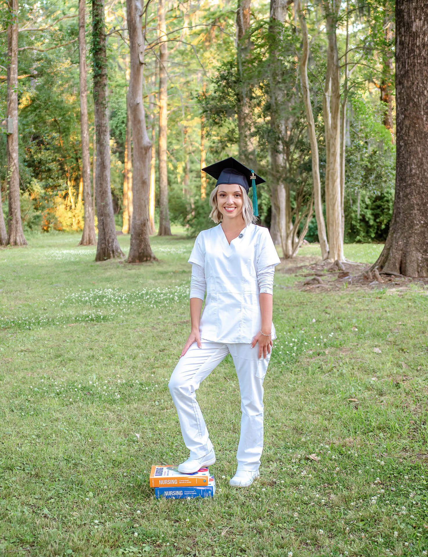 A UNCW senior in a cap and white nursing scrubs standing in a wooded area in Wilmington, North Carolina, with textbooks at her feet. This image captures a blend of academic and career aspirations, perfect for capturing meaningful senior photos.