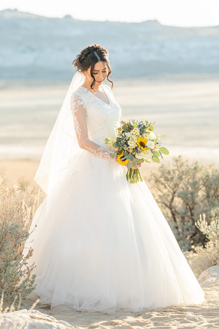A bride wearing a white wedding dress with long lacy sleeves holds a bouquet of sunflowers at Antelope island in early spring. Captured by Salt Lake Photographer Melissa Woodruff