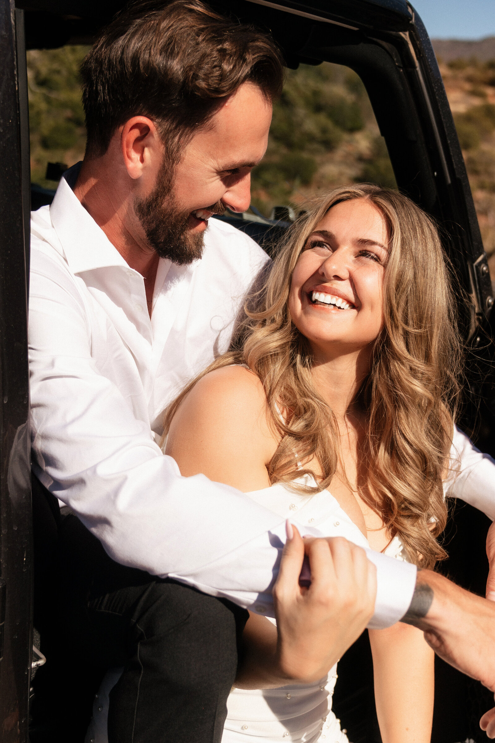Bride and groom laughing together in the back of a black Jeep during their adventurous elopement at Merry Go Round Rock in Sedona, Arizona, wearing white and surrounded by desert scenery.
