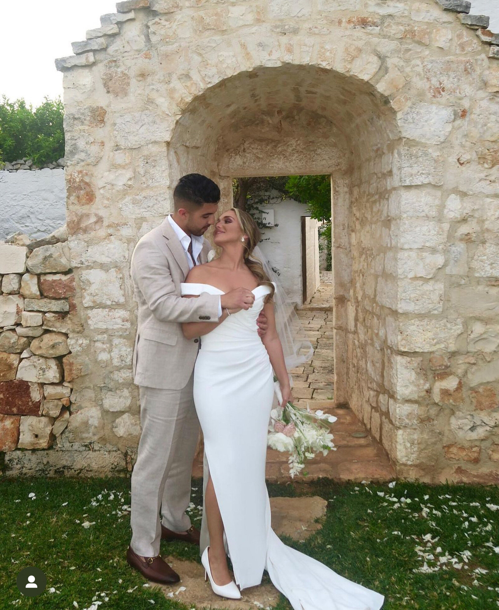 bride in a white off-shoulder gown holding a bouquet of flowers stands under an archway, gazing upwards at the groom in a light beige suit