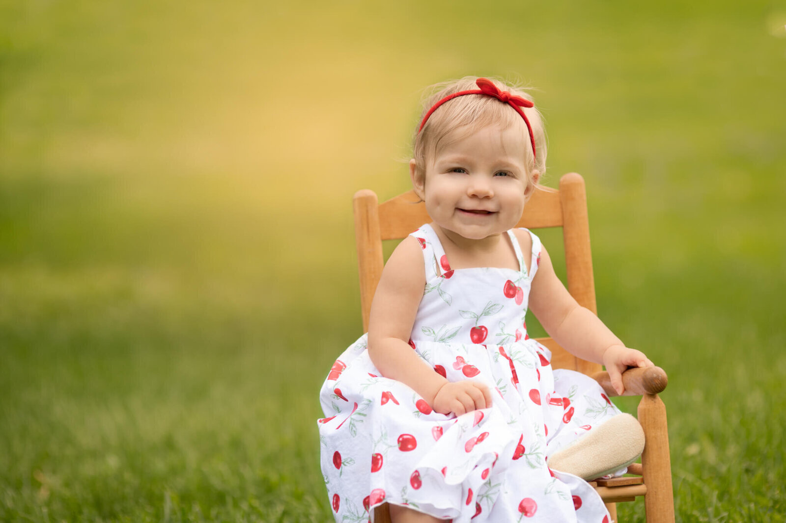 e year old girl wearing white dress with red cherries and a red hair bow sitting in a chair outdoors