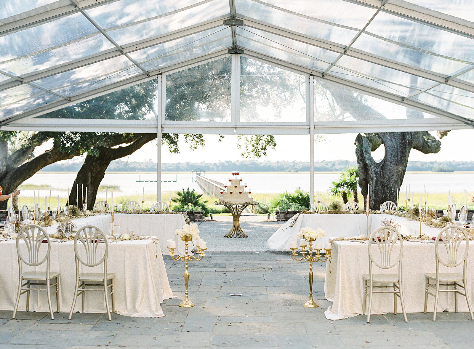 Reception at Lowndes Grove under glass tent looking out to the oaks, dock and water. Long tables forming an open square. Gold chairs, tall floor gold candelabras with white pillar candles. Gold painted myrtle leaves and gold painted baby's breath running down middle of all tables and cascading down the ends of the tables. Gold candlesticks with white tapper candles. Drink table in the middle behind the tables with half white drink and red drink in champagne glasses. Photographed at Lowndes Grove wedding by wedding photographers in Charleston Amy Mulder Photography.