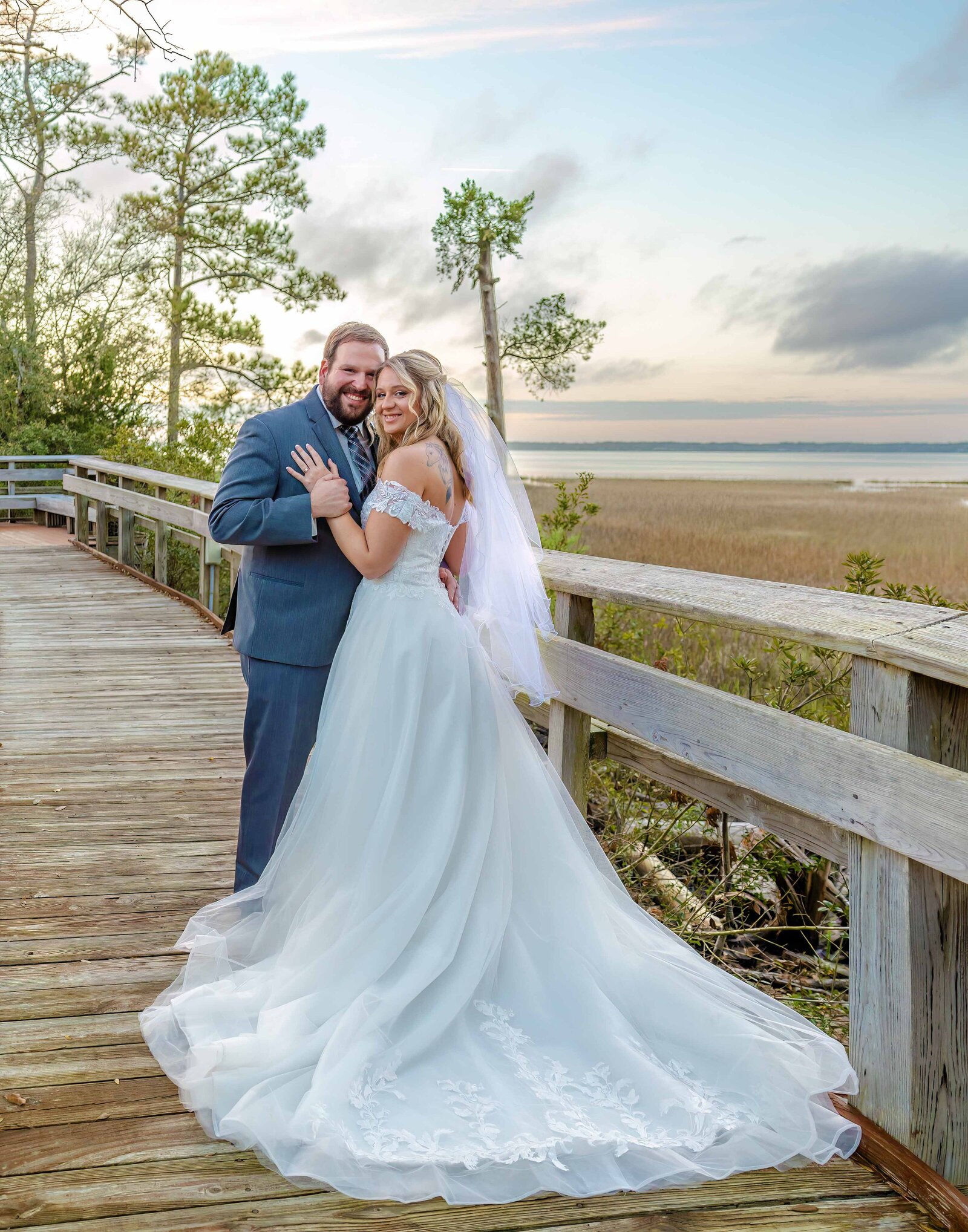 The bride and groom pose together on a wooden boardwalk overlooking the marsh at sunset, captured during a Pine Knoll Shores Aquarium wedding, blending natural beauty with romantic charm.