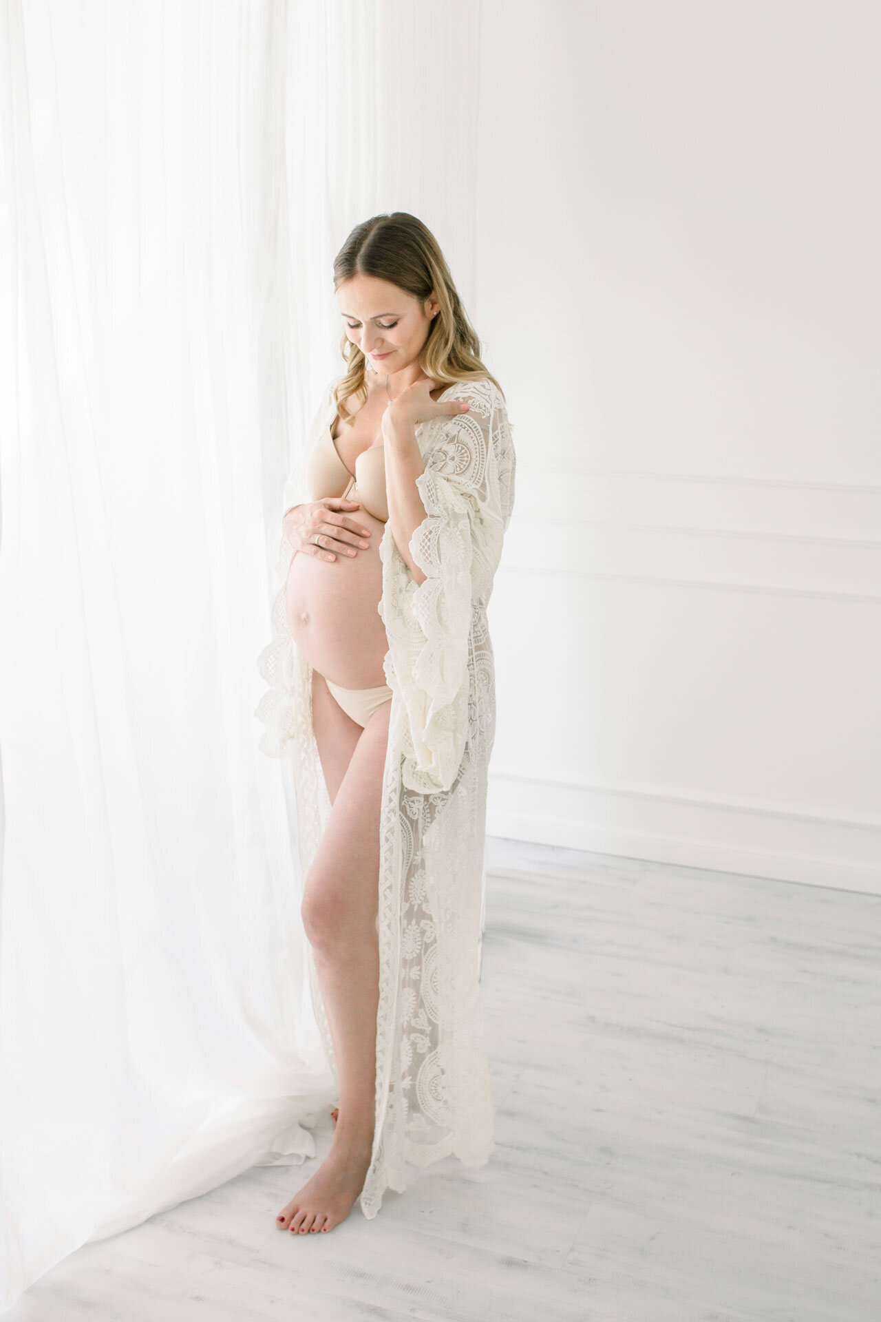 Schwangerschaftsfotografie im Fotostudio Gütersloh bei Bielefeld. Schwangere vor heller Kulisse und weißer Gardine in Dessous und langem Kimono aus Spitze.