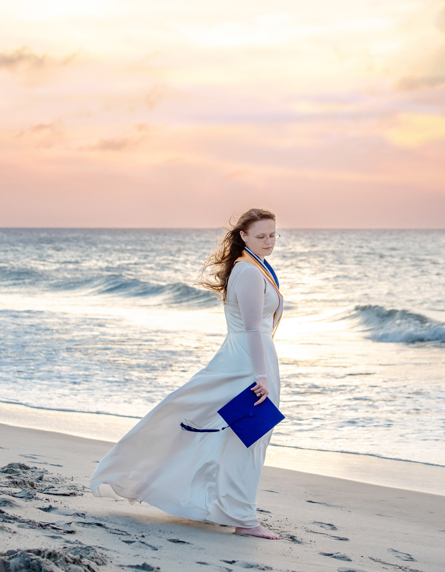 A CFCC senior in a flowing white dress holding her cap with tassle blowing in the wind, walking along Carolina Beach at sunset in North Carolina. This elegant image highlights the celebration of graduation, perfect for capturing memorable senior photos by the ocean.
