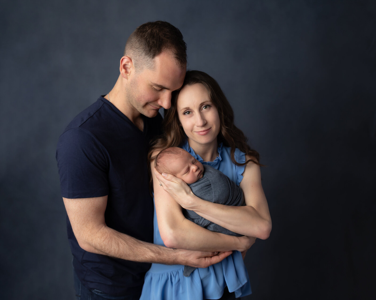mom and dad holding newborn baby boy wrapped in blue for newborn studio portraits