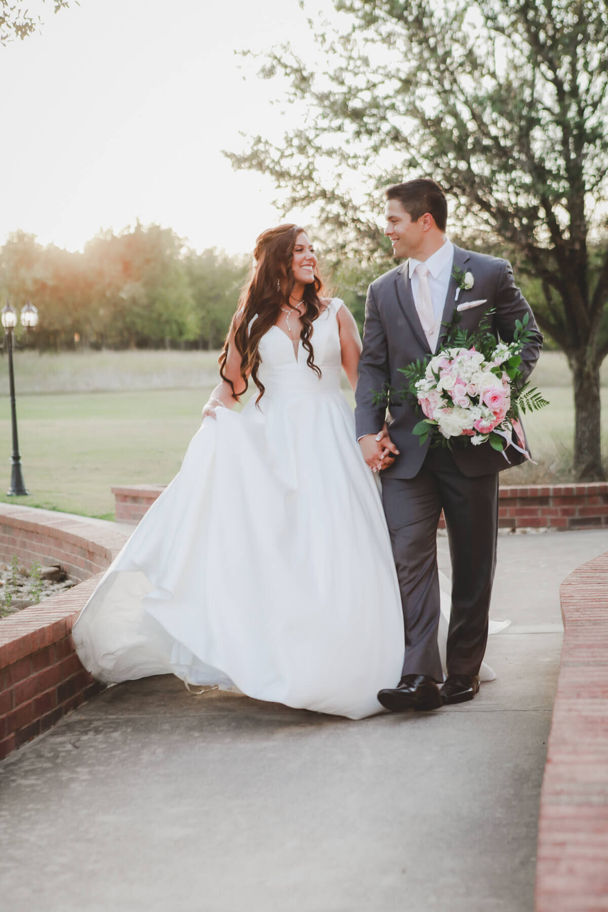 A bride and groom walk hand in hand by a brick pathway.