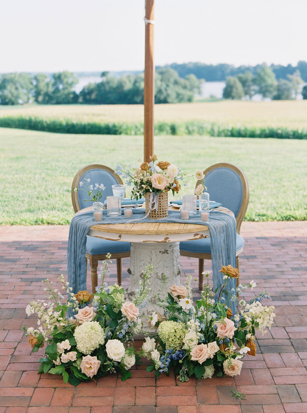 Sweetheart table with bridal bouquet, candles and ground arrangements in front of it with florals to include green hydrangea, blue delphinium, toffee roses, blush garden roses, and white cosmos.