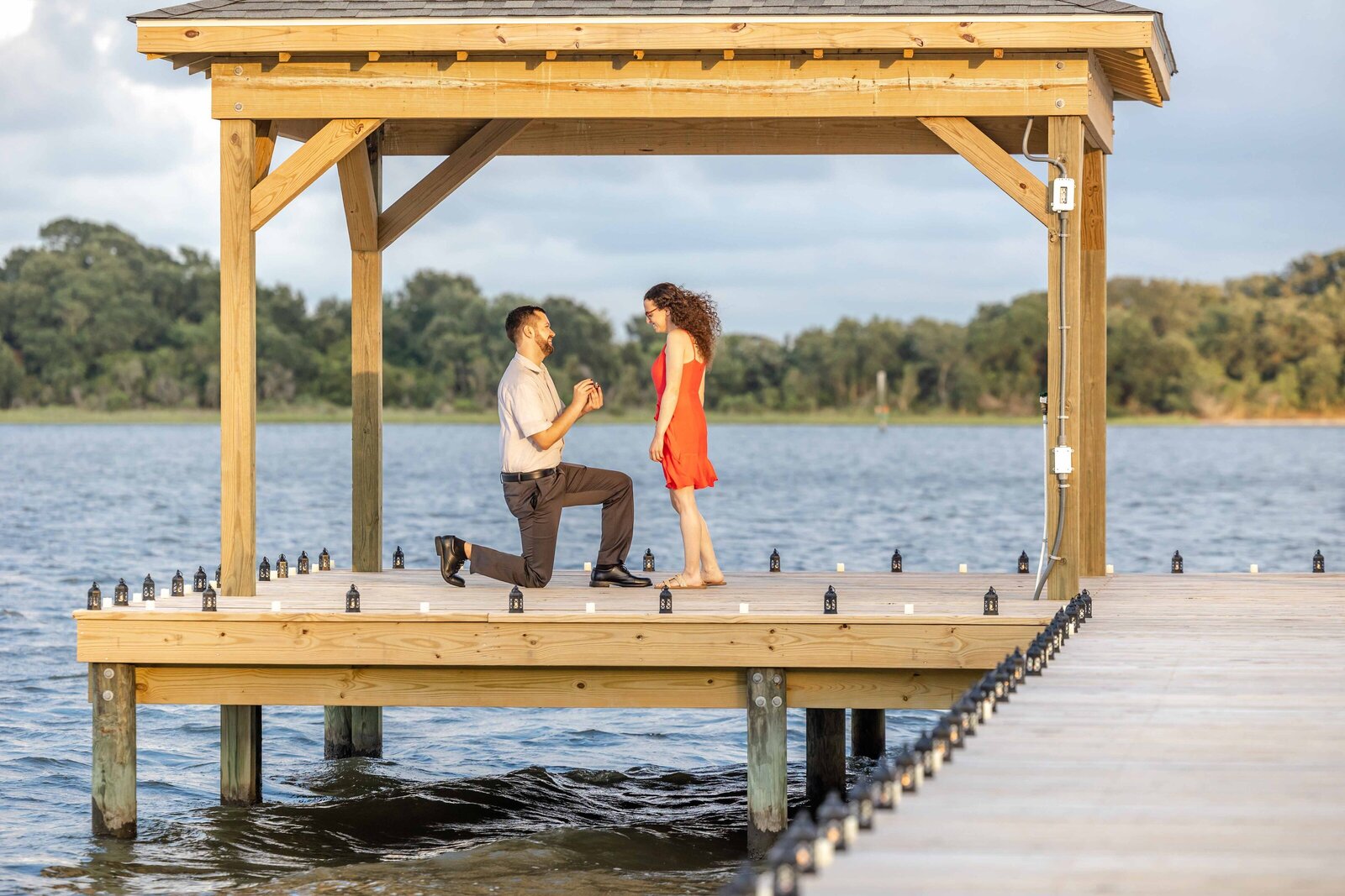 A waterfront proposal as a man kneels on a wooden dock under a gazebo, holding a ring for the woman in a bright red dress. The calm water and surrounding greenery create a romantic and serene atmosphere, perfect for North Carolina engagement photography.  