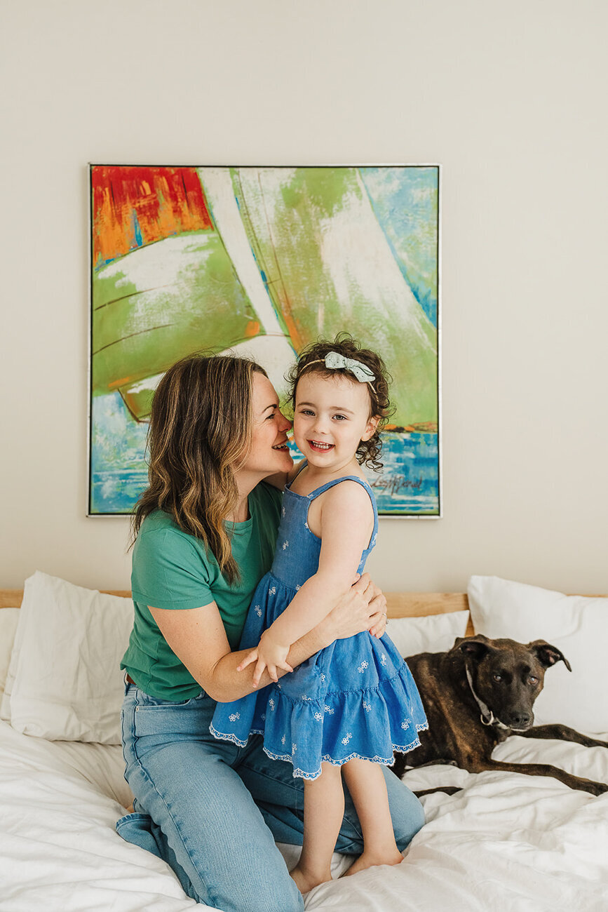 mom snuggles daughter on white bed with colorful artwork behind