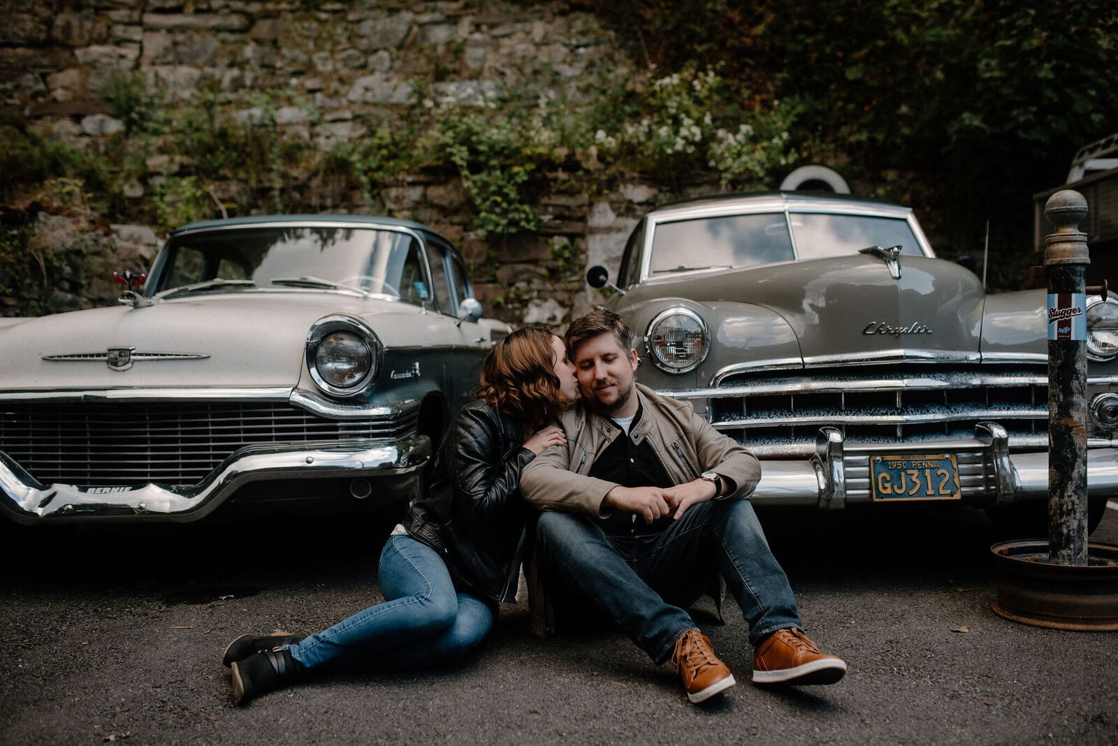 couple sitting in parking lot in font of antique cars whispering into ear