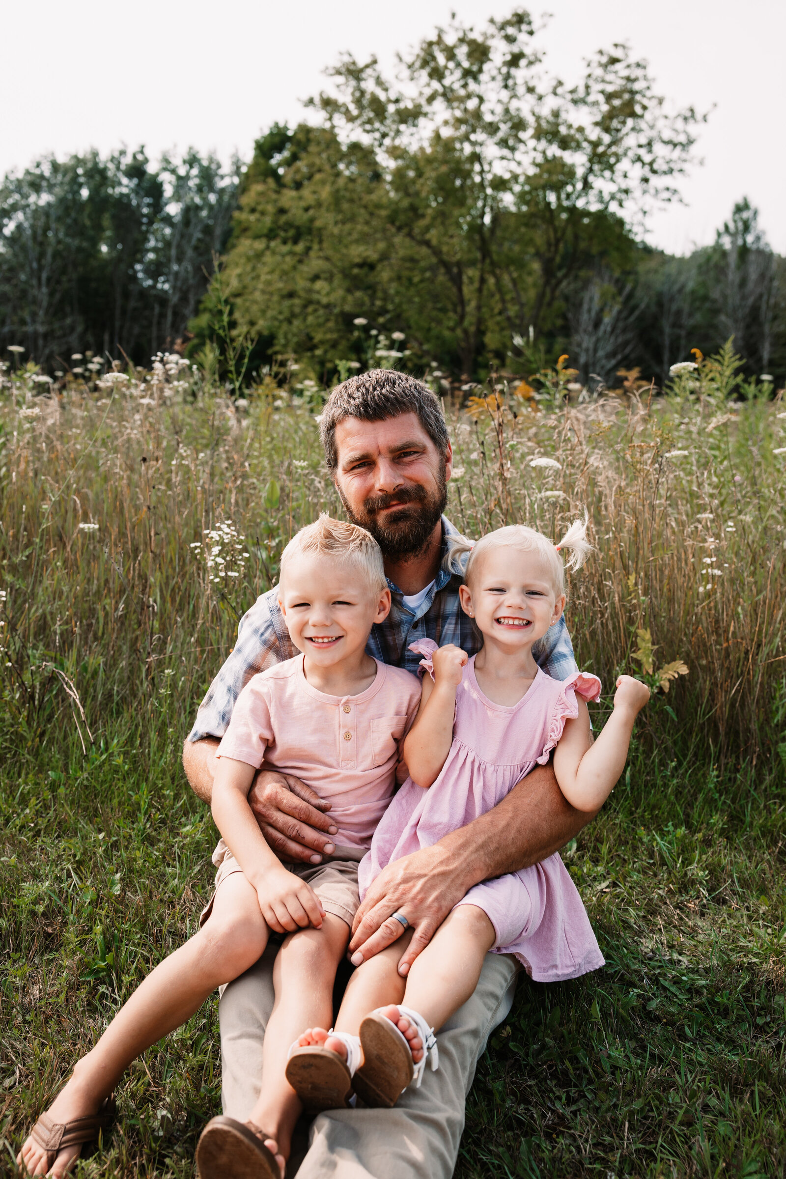 A happy father sits in a field of wildflowers with his toddler son and daughter in his lap