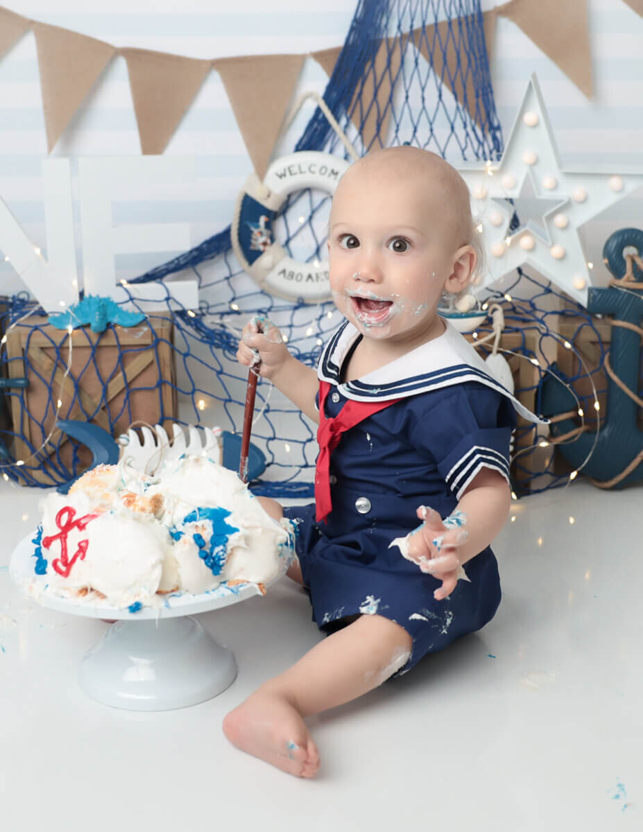 A smiley boy celebrating his birthday with a nautical-themed cake smash in a Rochester, NY studio. Colorful decorations featuring anchors and sailboats surround him as he playfully digs into a beautifully decorated cake.