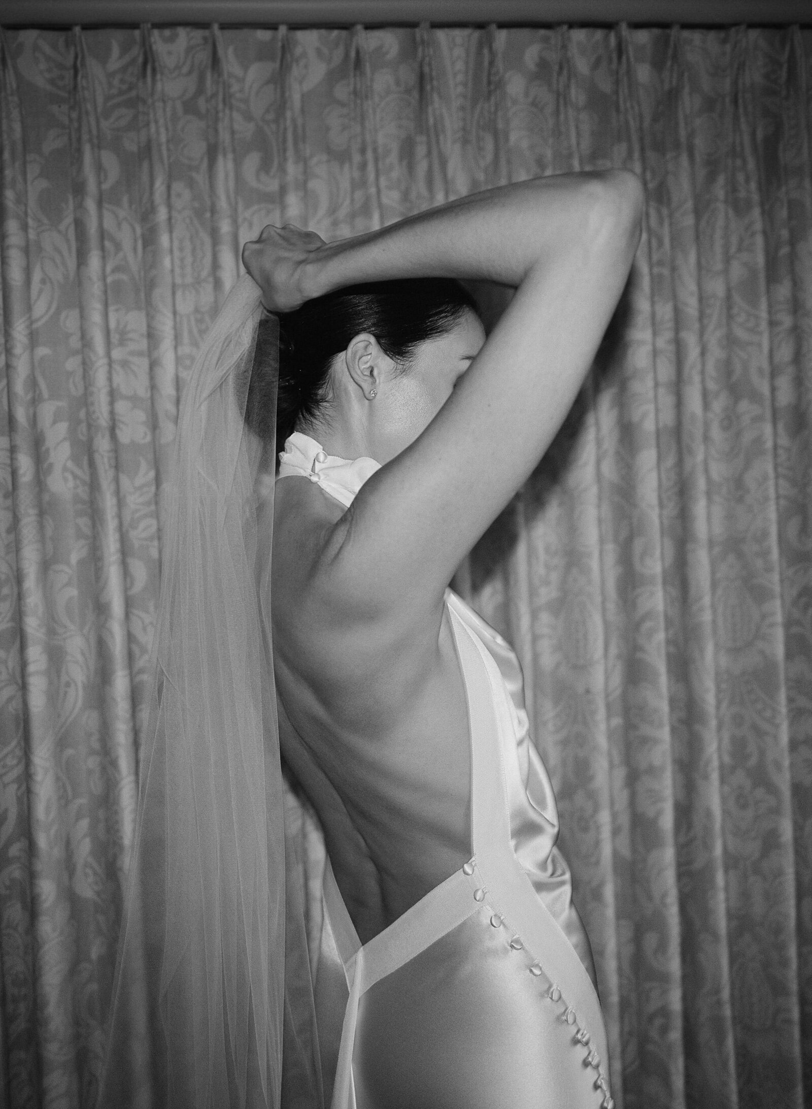 Candid direct flash black & white photo of bride in backless silk dress fixing her veil in front of floral curtains 