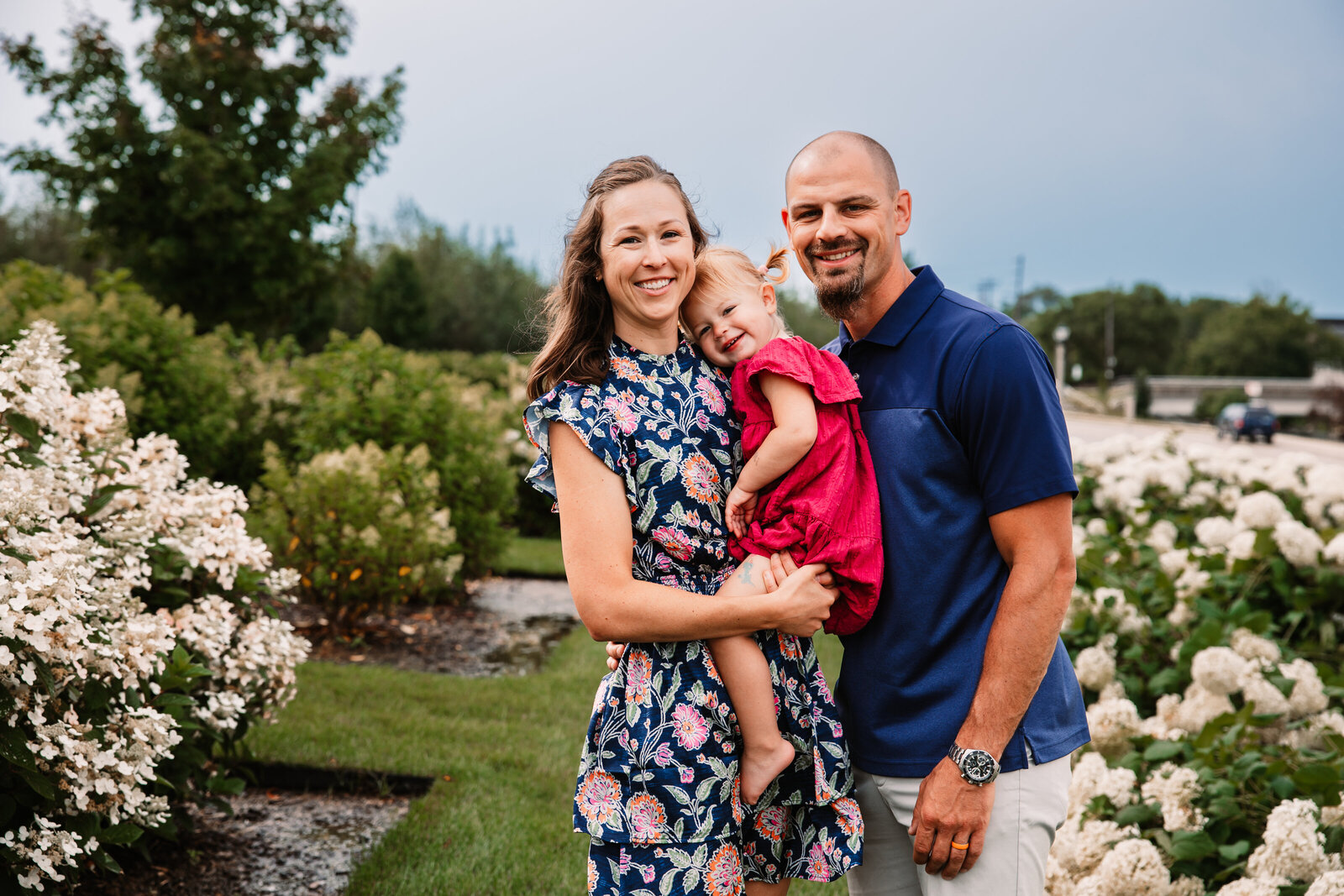 A mom and dad carry their toddler daughter through a park garden of white flowers