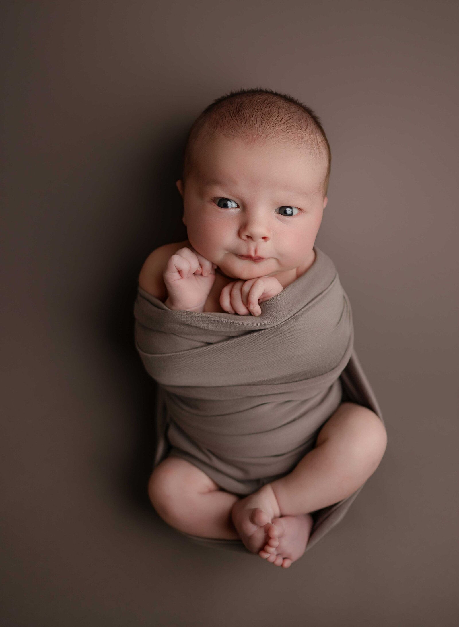 Baby boy in brown swaddle, looking at camera awake.