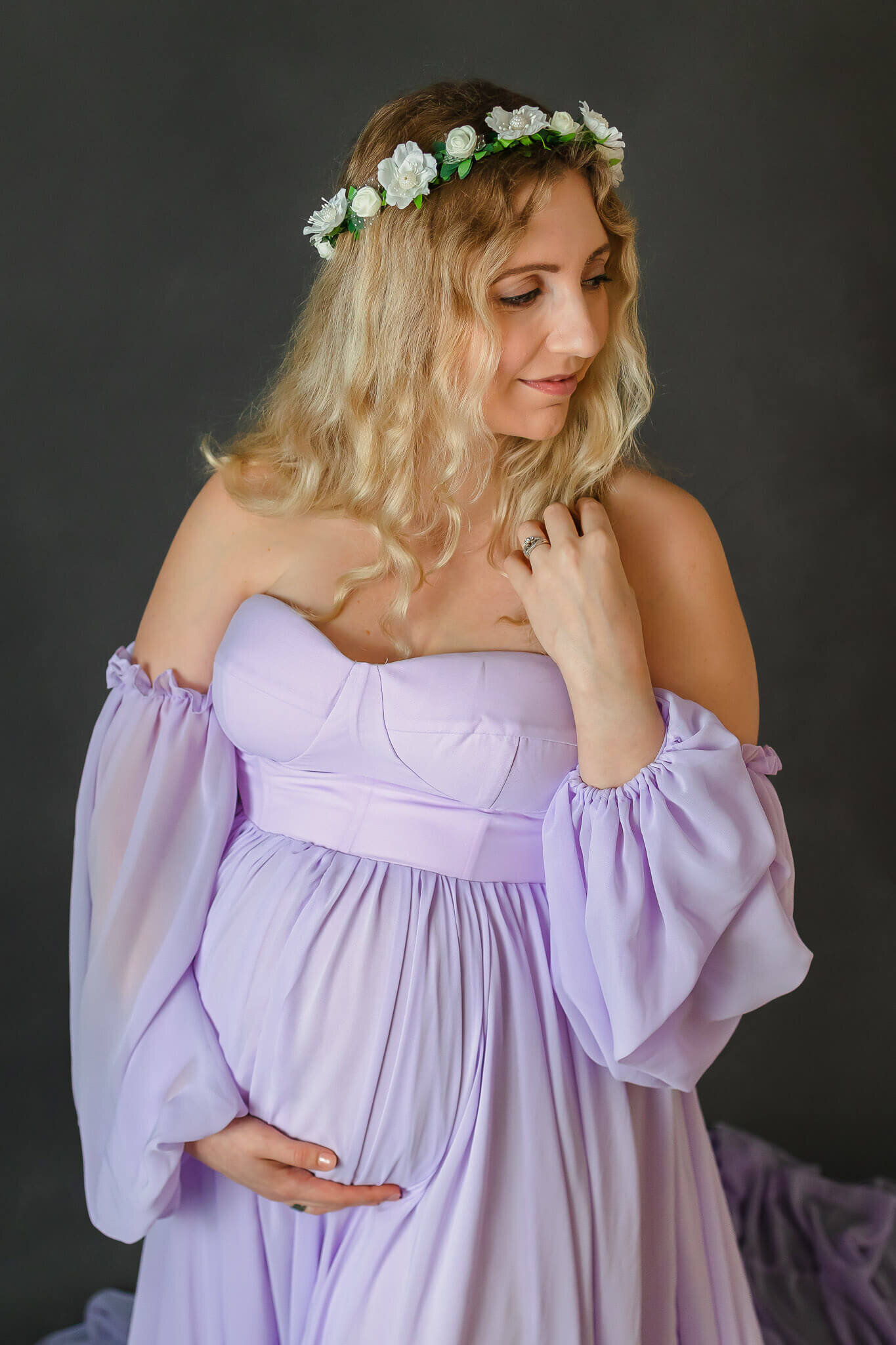 An expecting woman in a purple dress holding her belly with one hand and playing with her hair with the other hand.