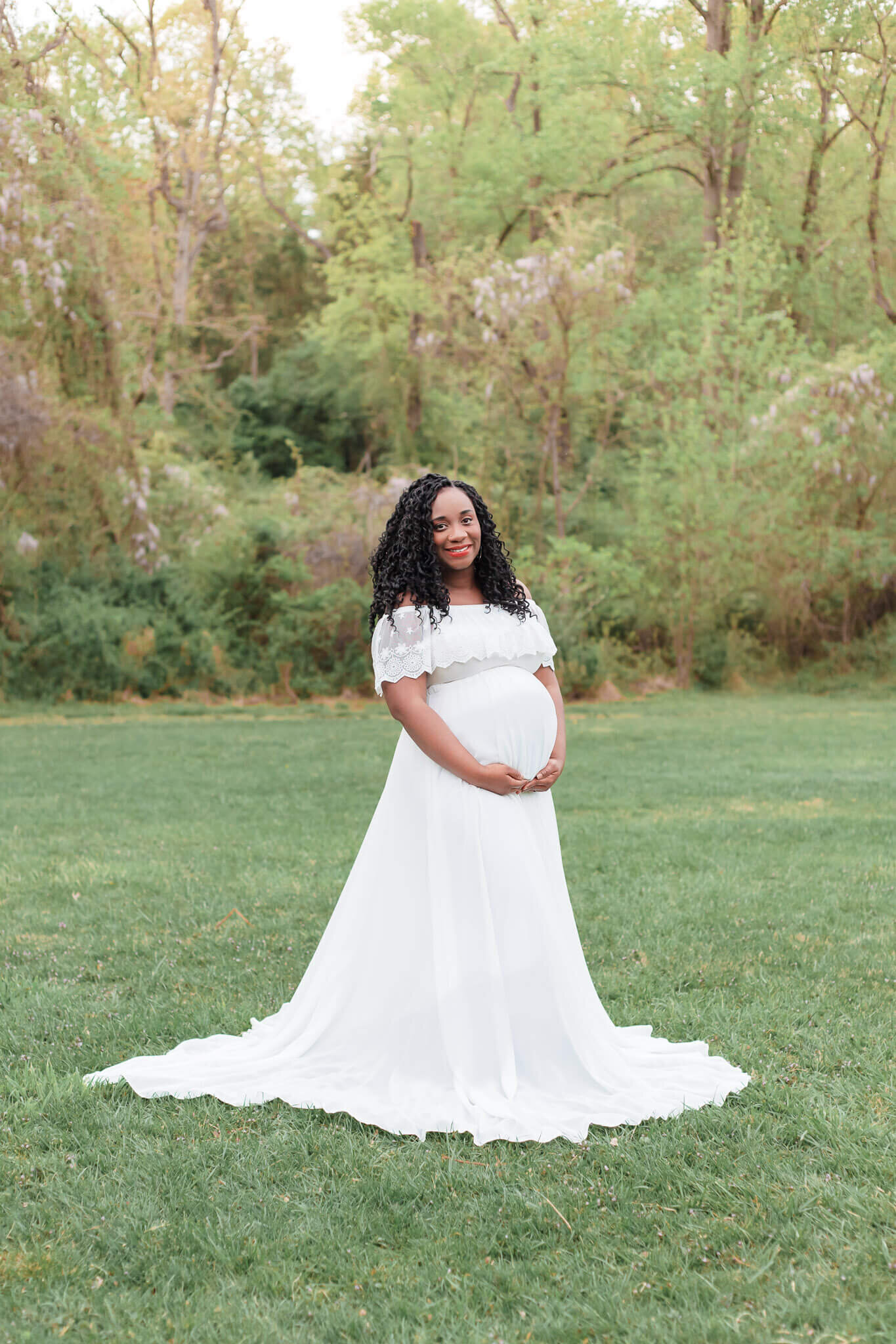 An expecting woman in a white dress hugging her belly in a field at sunset.