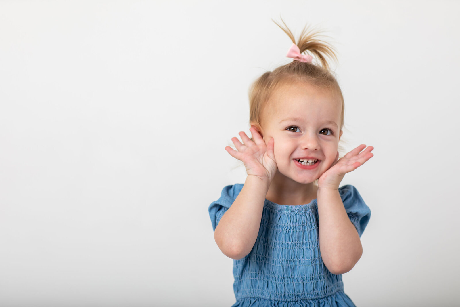 Adorable toddler girl in a blue outfit squeezes her cheeks with both hands, making a playful and silly face