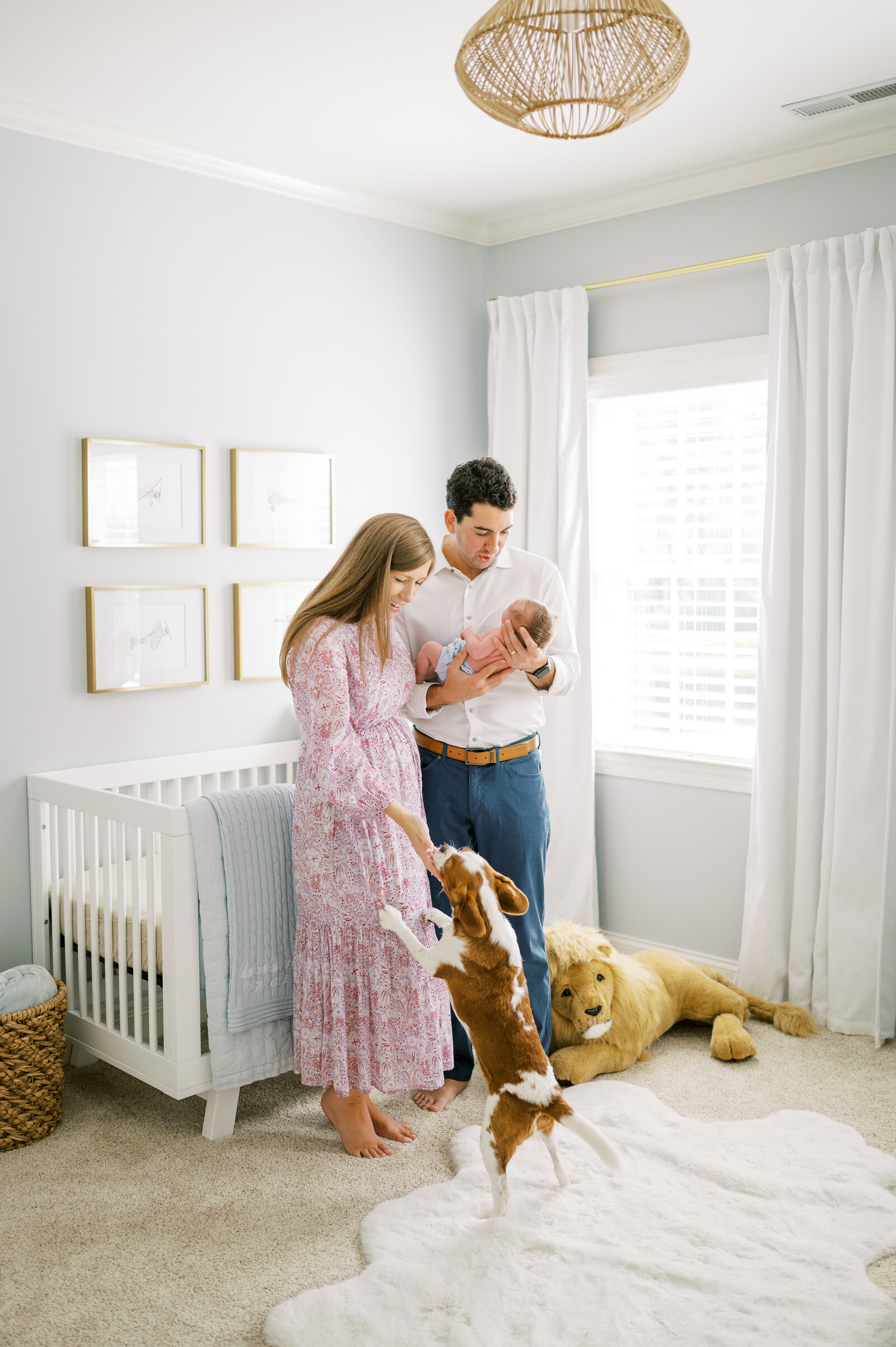 Man and woman stand in front of baby crib with Cavalier King Charles dog and newborn baby during photo session