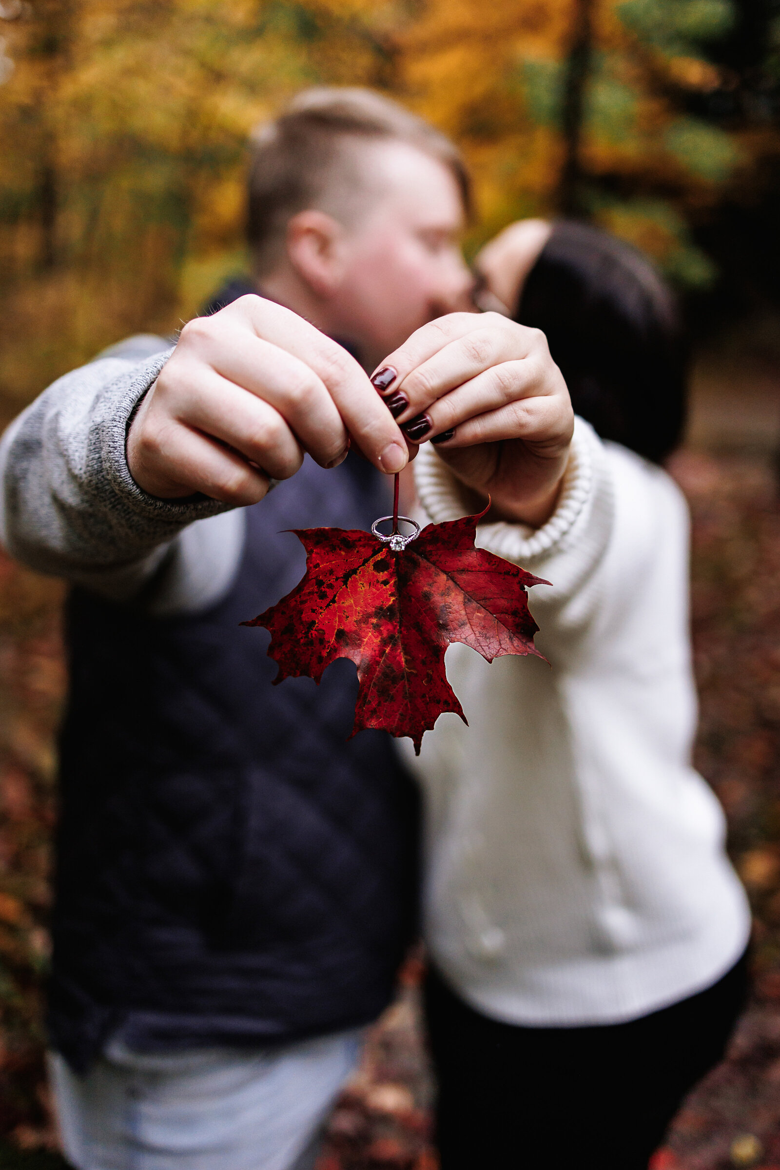 Couple holds out a fall leaf with her ring  placed on the ste,m as they share a kiss