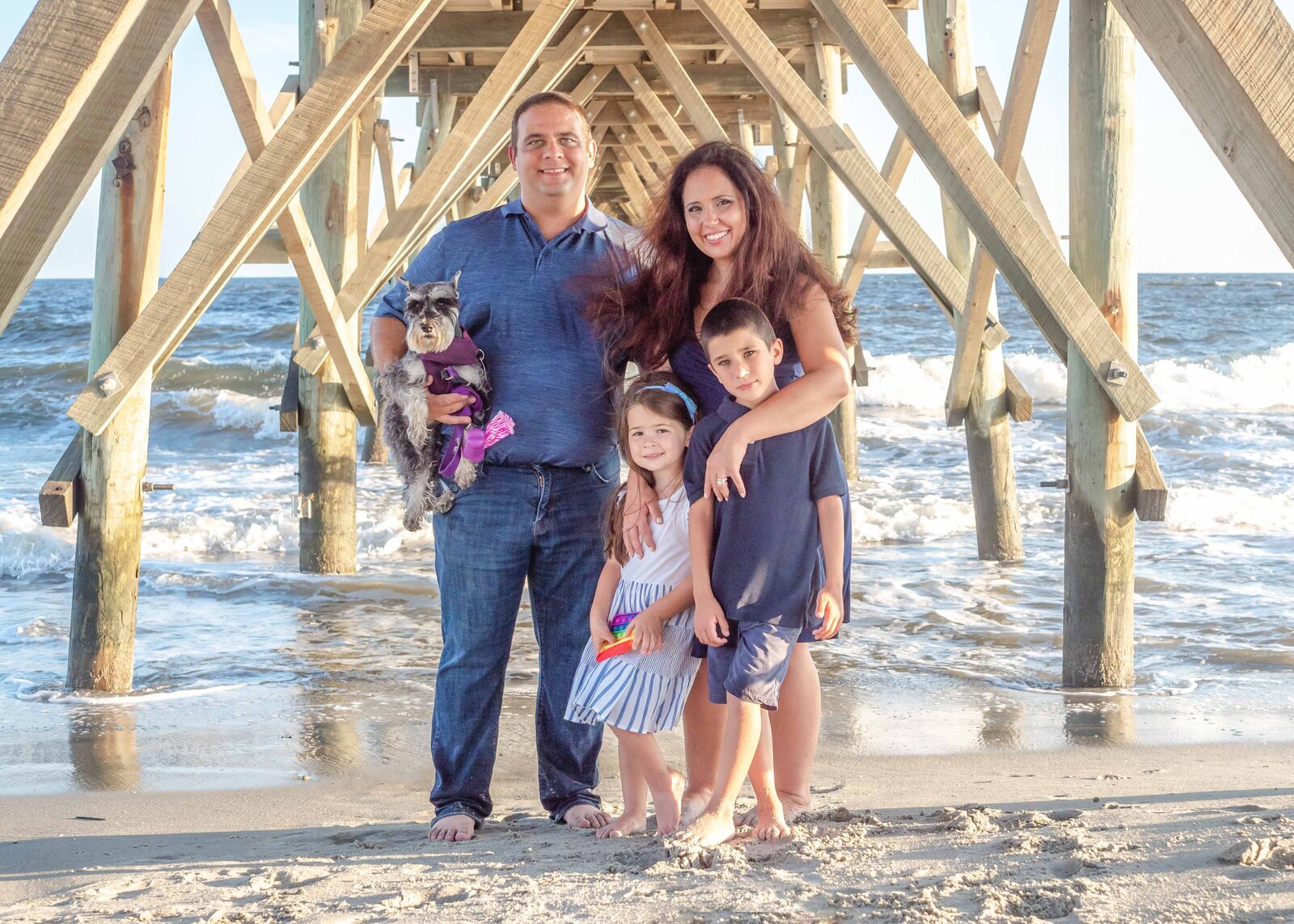 A family of four poses with their dog under a wooden pier, dressed in navy and striped outfits. The ocean waves and pier structure provide a dynamic backdrop. Ideal for artistic family photography in Wilmington, NC, showcasing fun and lively beach moments.