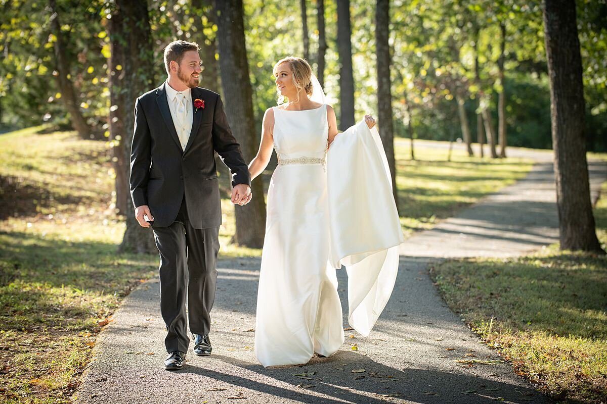 the groom wearing a black suit guides the bride, wearing a long white satin gown by the hand down a garden path