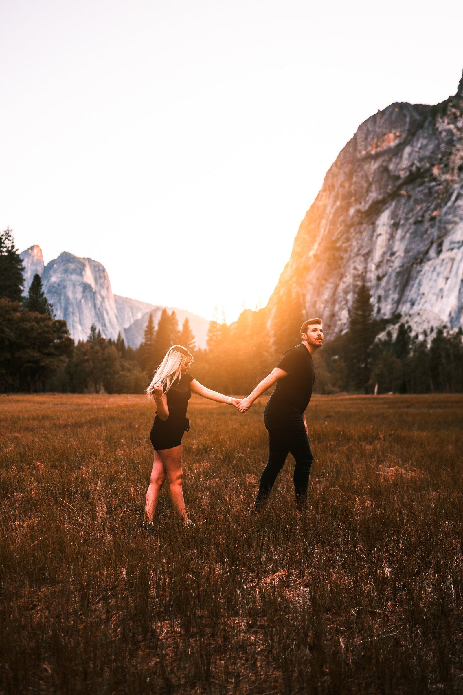 Couple holding hands and walking through field with sun setting behind them.