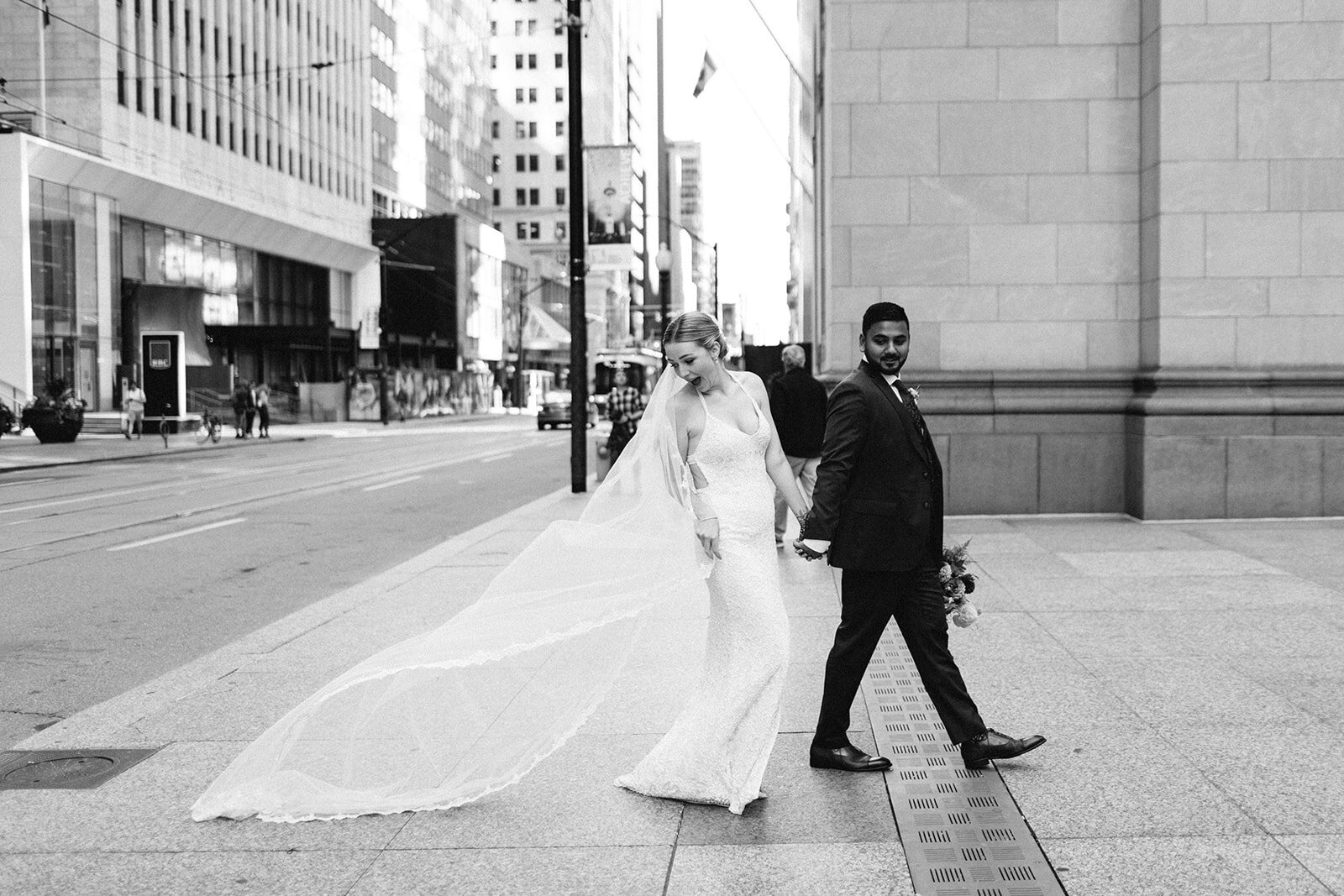 Bride and groom holding hands, walking across an urban street in a beautiful black and white image captured by Toronto destination wedding photographer, Jennifer van Son, at Steam Whistle Brewing. The bride's veil flows elegantly behind her, encapsulating a moment of timeless romance.