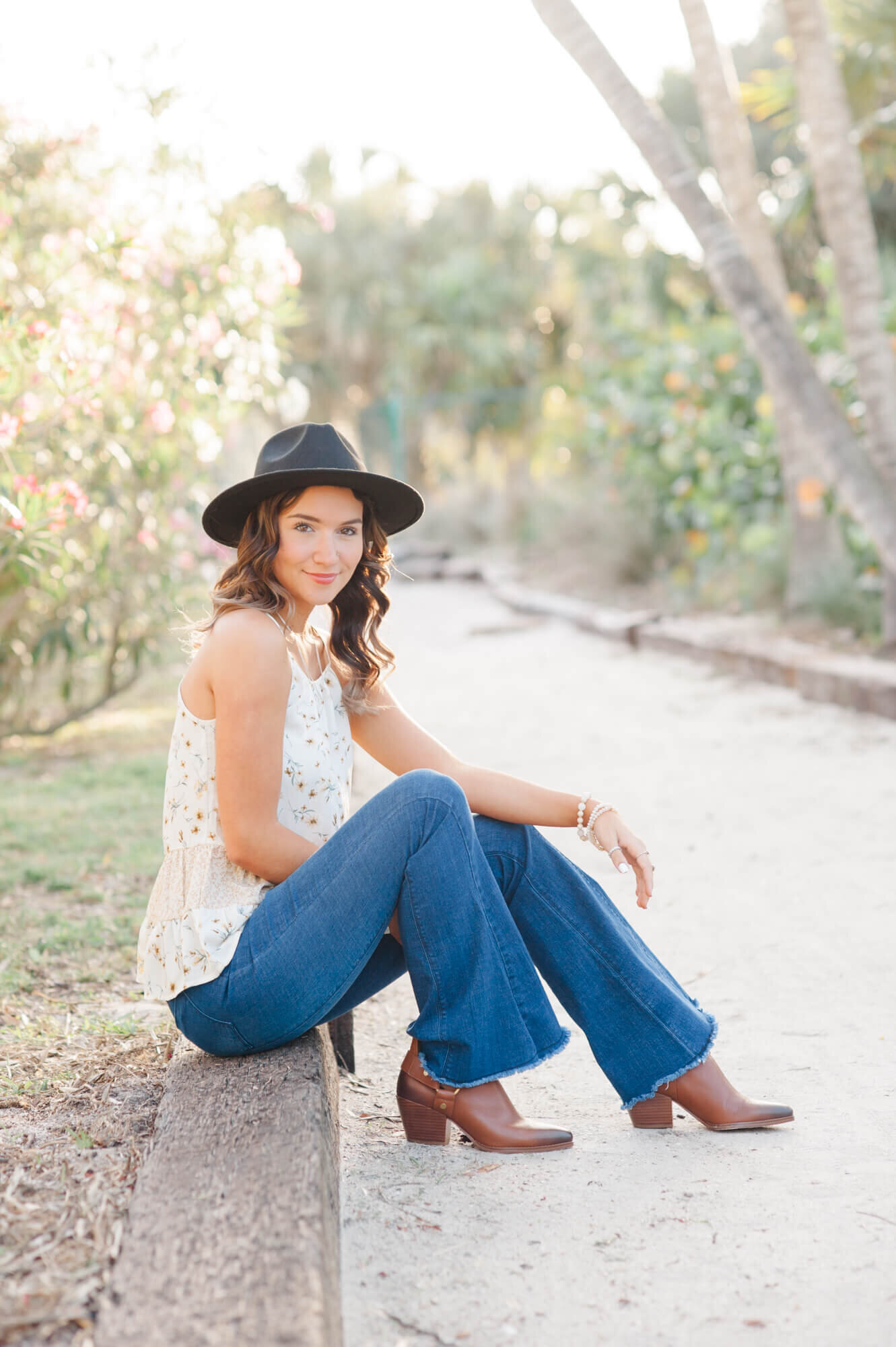 Senior girl wearing a wide brim hat and boots sits on a beach path during her senior photoshoot