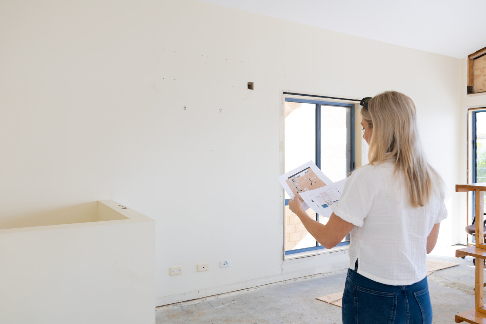 A woman examines renovation plans in a partially finished room with white walls and exposed flooring, embodying the bespoke style of a Newcastle Kitchen and Bathroom Designer.