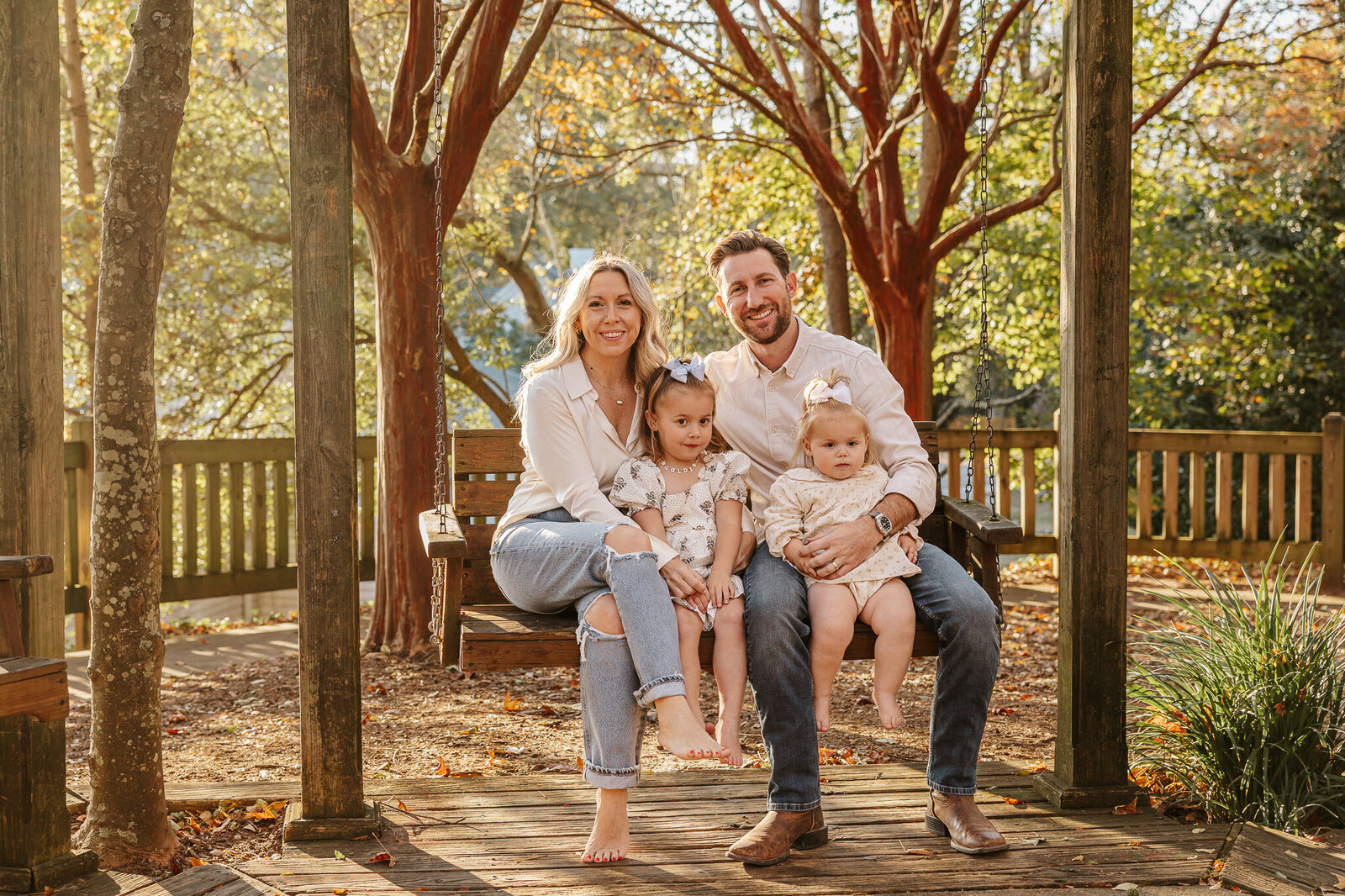 Family session at Hopelands Gardens in Aiken SC - parents sitting on a wooden walkway with their two your children, smiling together under the trees.