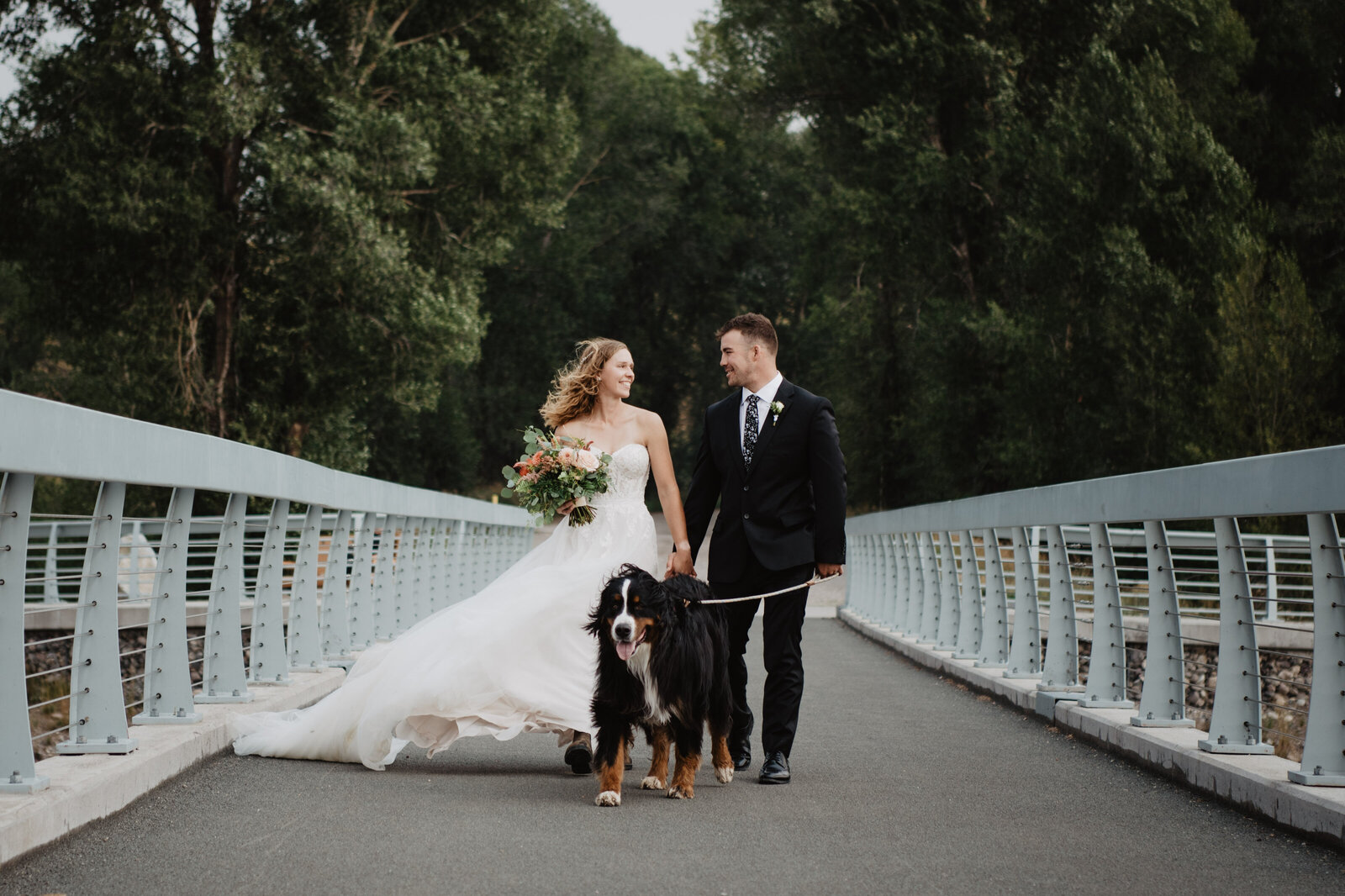 grand teton wedding location at Emily Stevens Park with bride adn groom walking their dog together on a bridge in the park over some water while the wind blows the brides gown