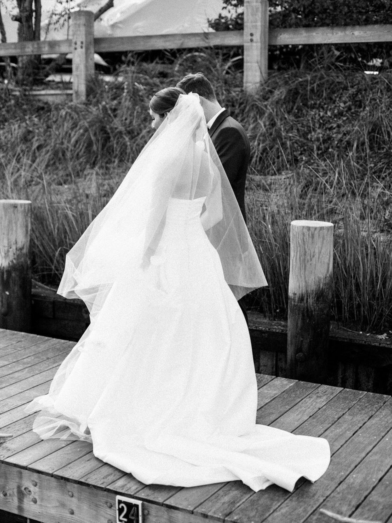 black and white photo of bride, wearing a flowing white gown and veil, walks hand-in-hand with the groom, who is dressed in a suit on a wooden dock with grass and a fence in the background